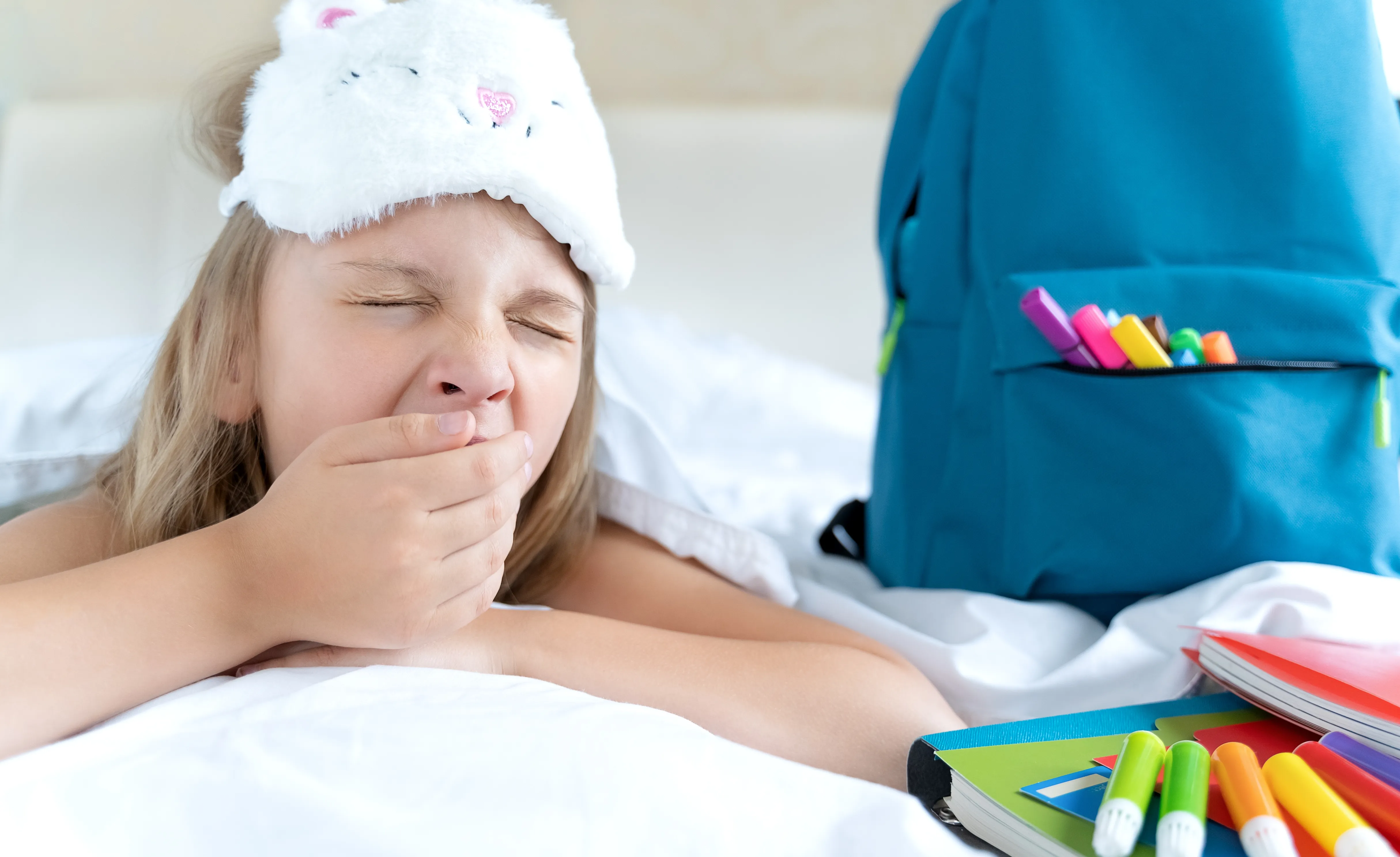 School-age child yawning in bed beside school supplies