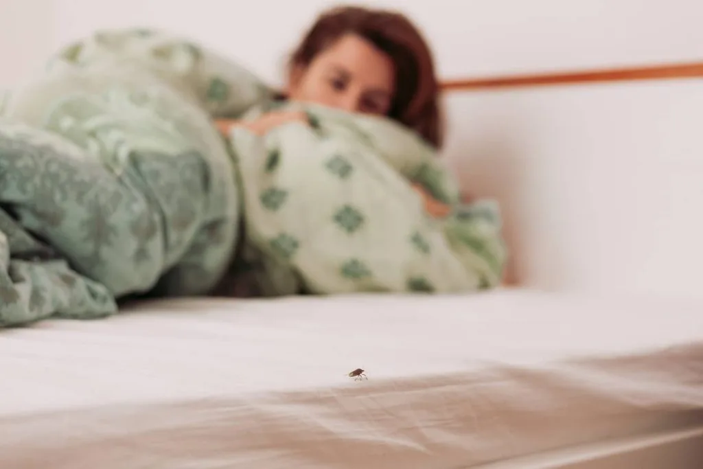A person is peeking from under a green floral blanket, focused on a single bed bug on the bedsheet.