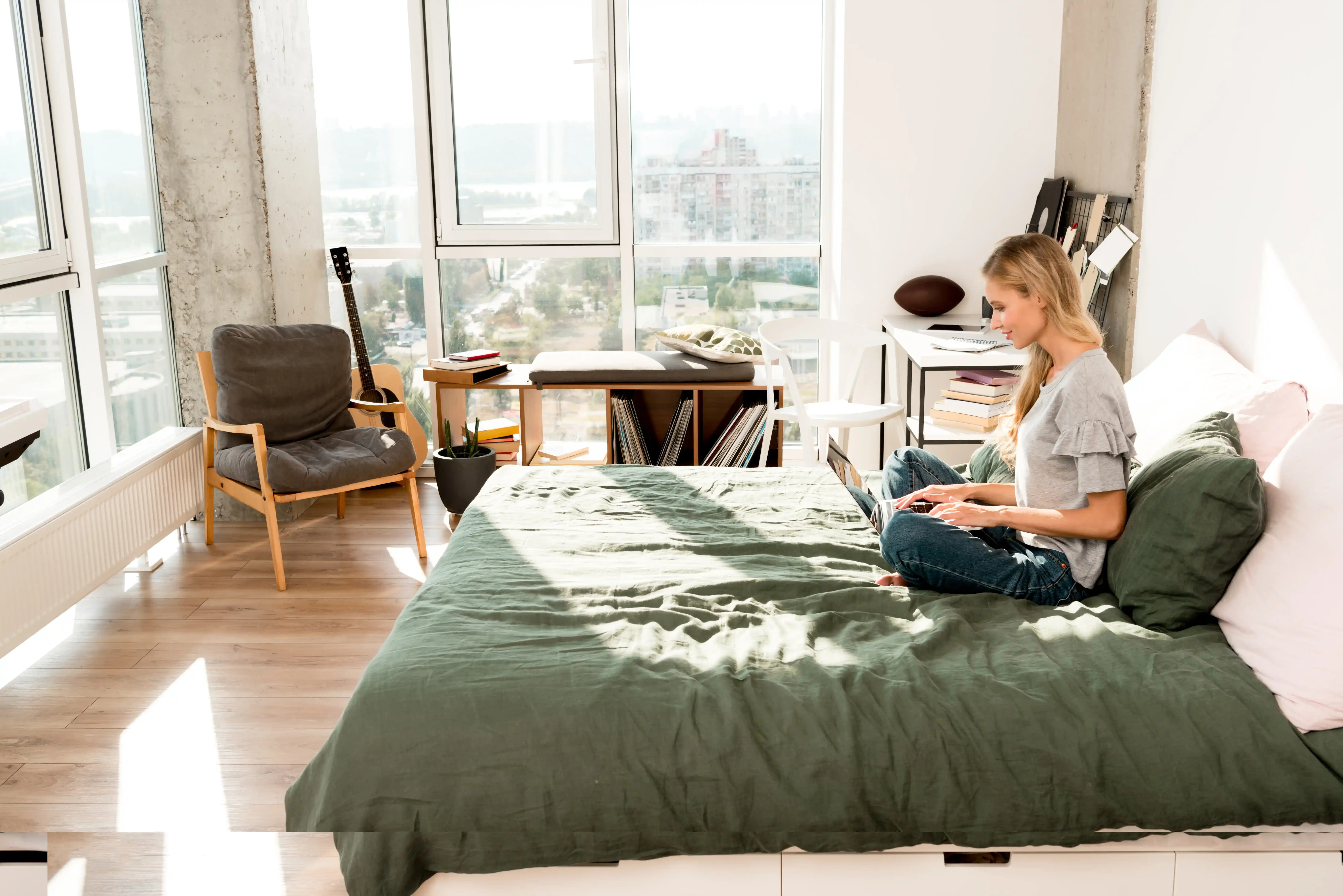  A person is sitting on a bed with a green duvet, using a laptop, in a bright room with a scenic city view through large windows.