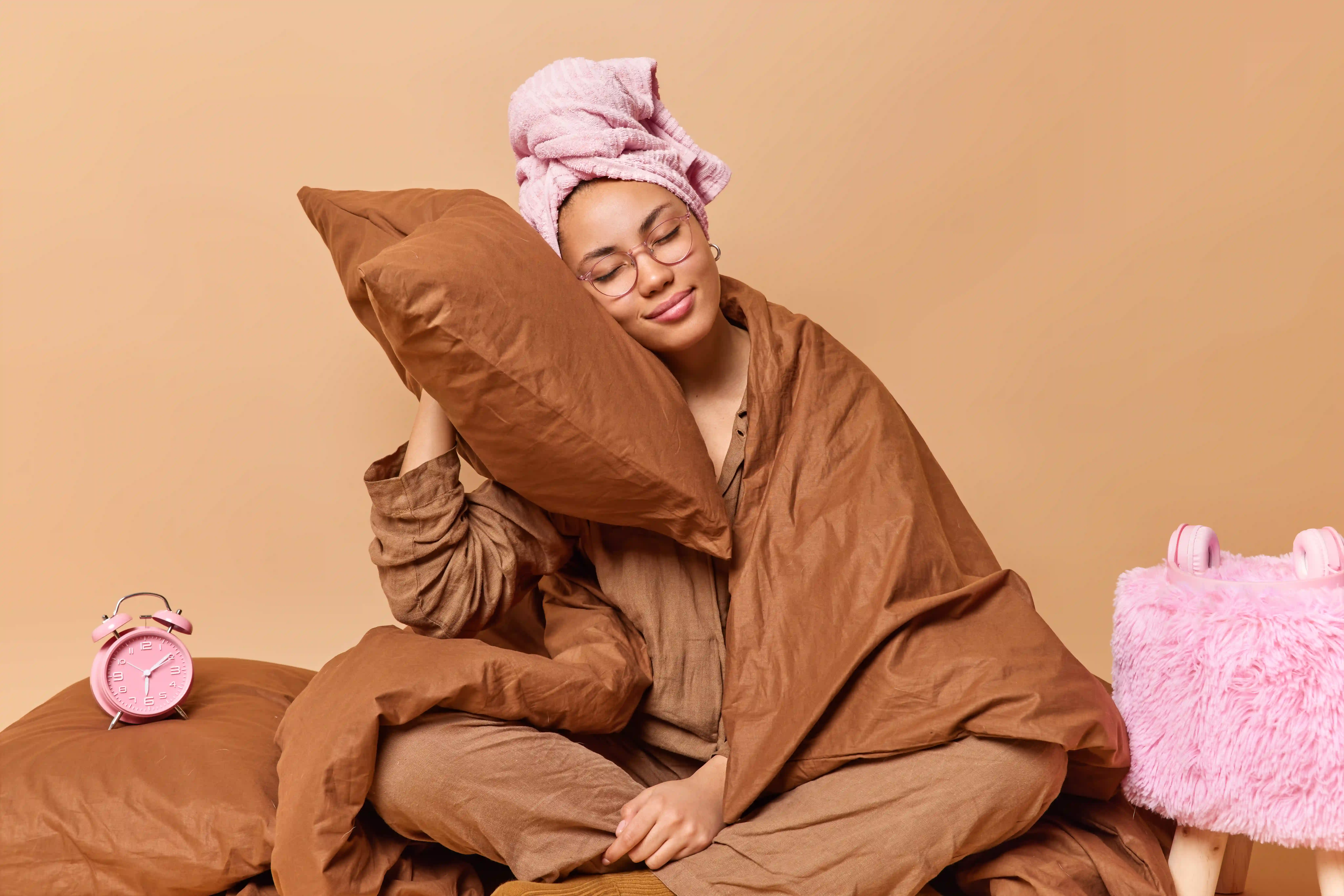 Blissful woman wears comfy nightwear leans head on soft pillow takes nap and daydreams wrapped in blanket wears towel wrapped on head sits crossed legs in bedroom isolated over beige background