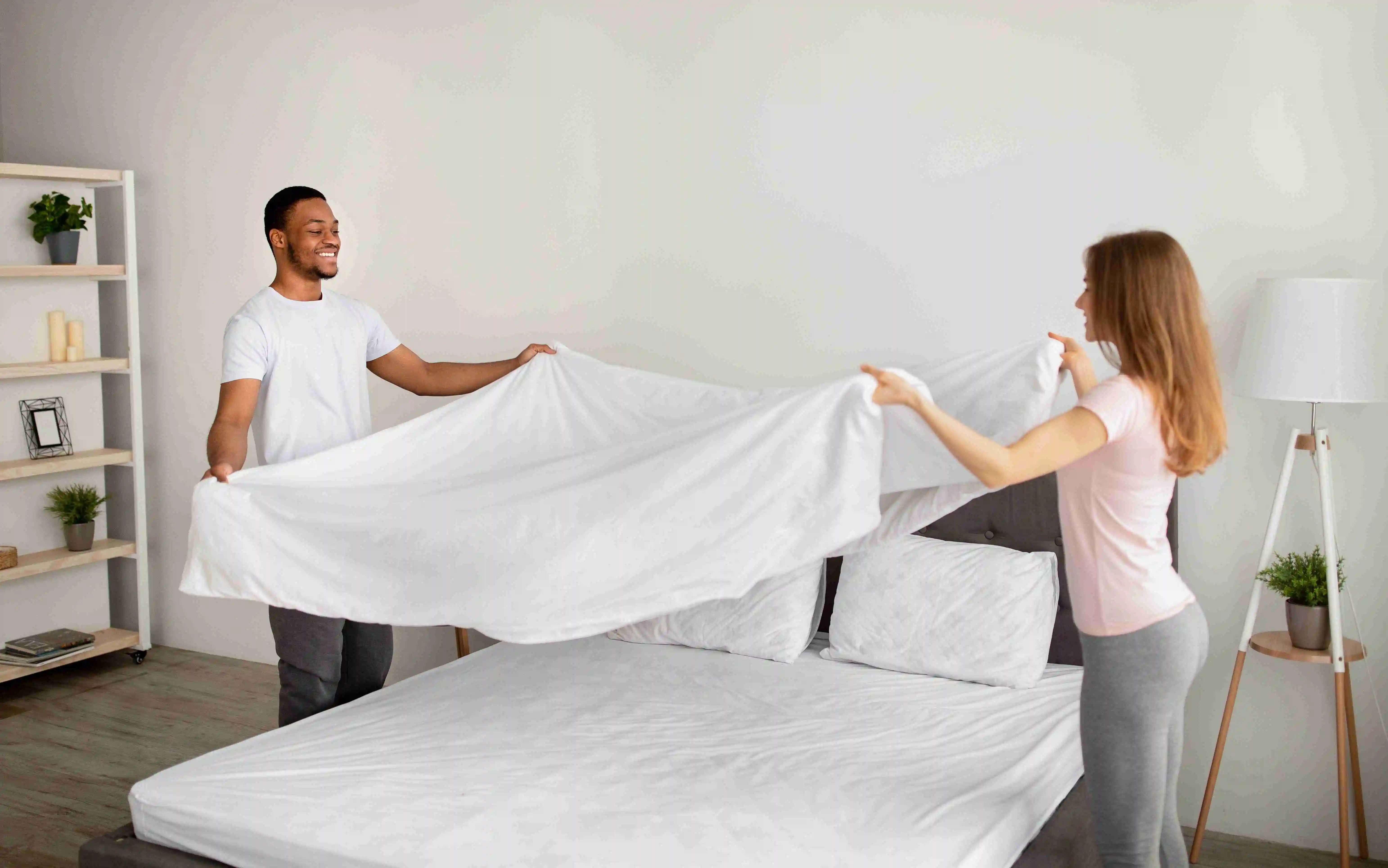 Cheerful multiracial couple making bed in morning, having fun at home