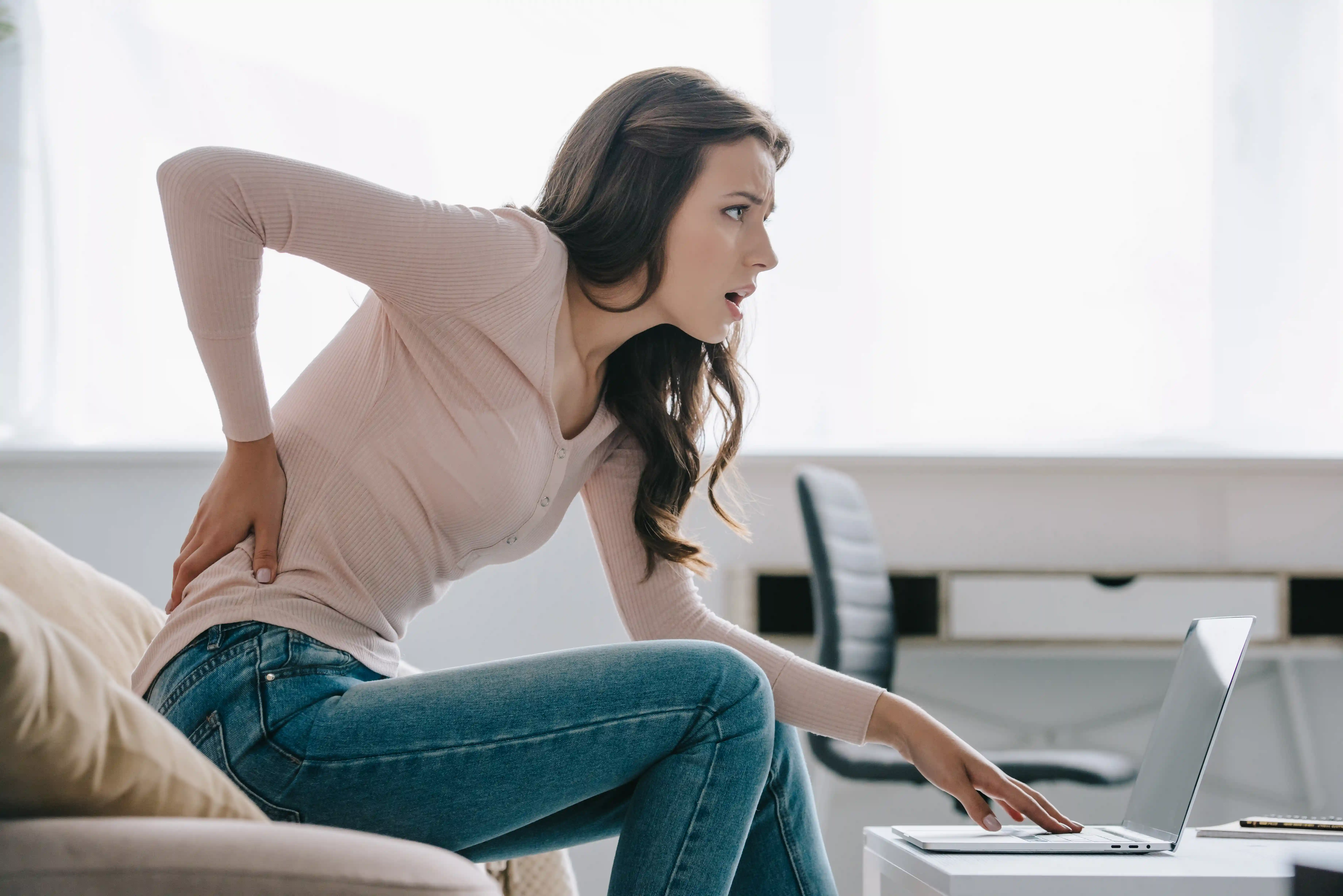 side view of young woman suffering from back pain while using laptop at home