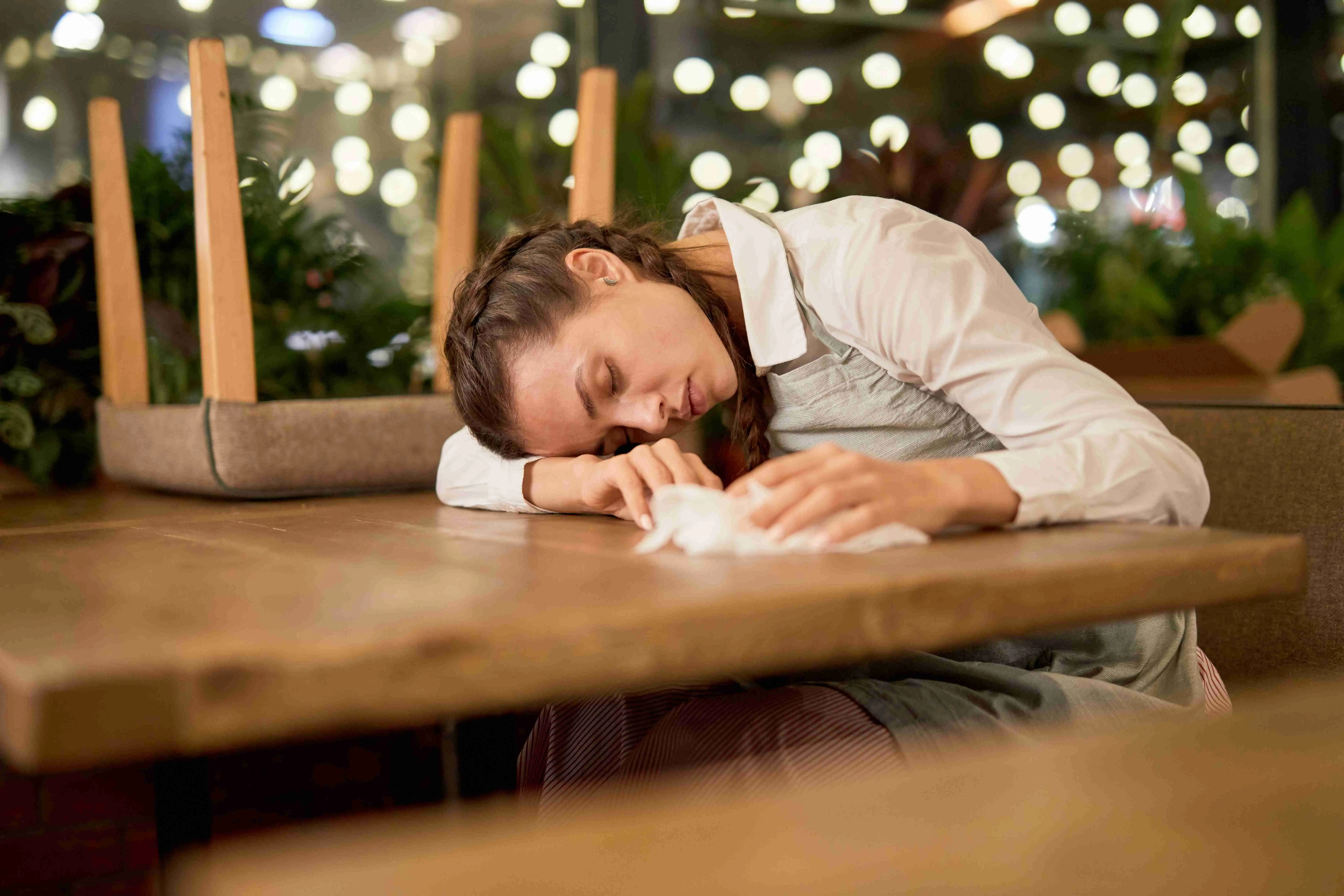 Tired waitress with duster sitting by table with her head on it and napping after work