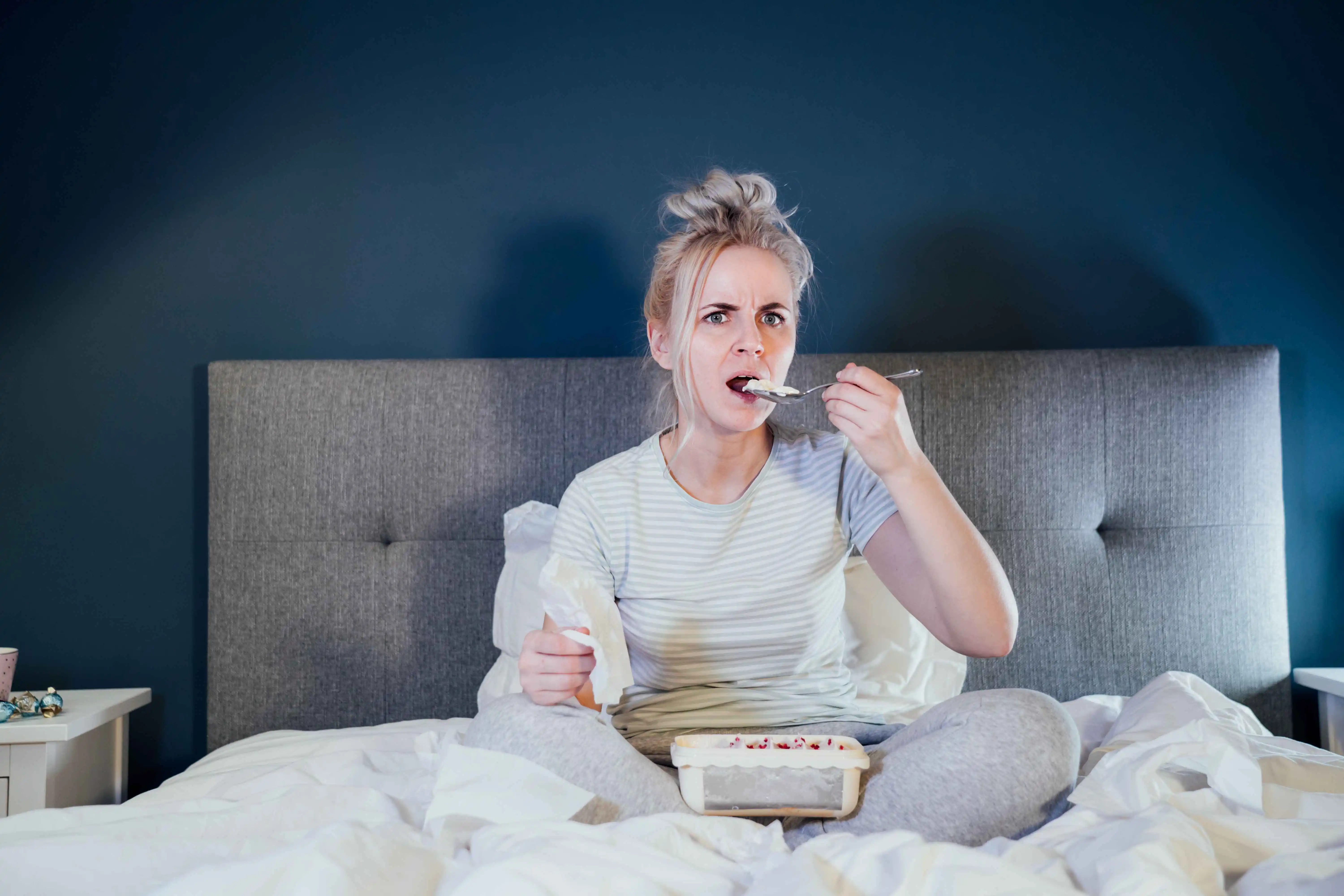 Shocked, stressed, surprised woman eating ice cream, holding a remote control while watching TV in bed.