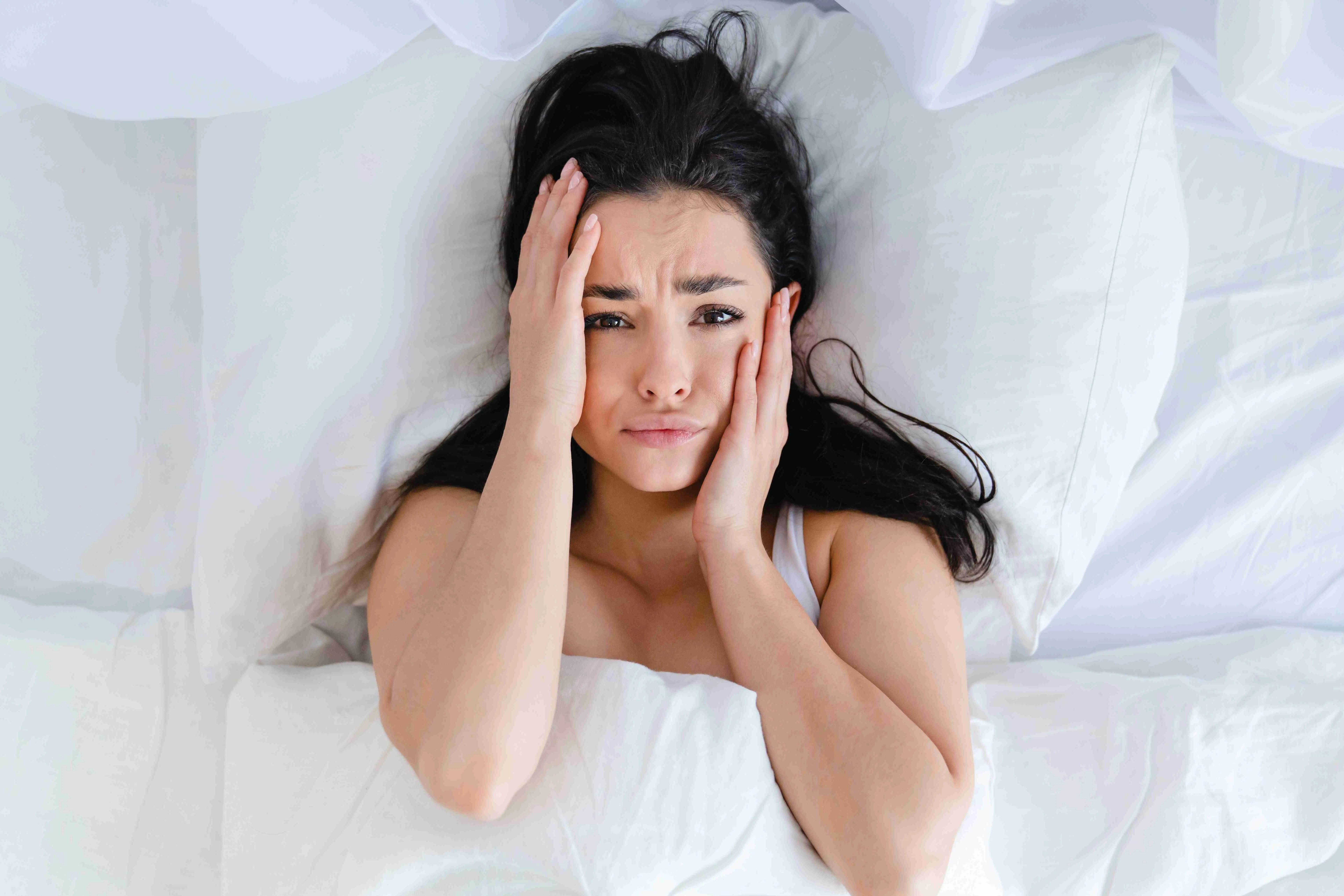 Sad young Caucasian woman lying in the bed with white linen because of the insomnia