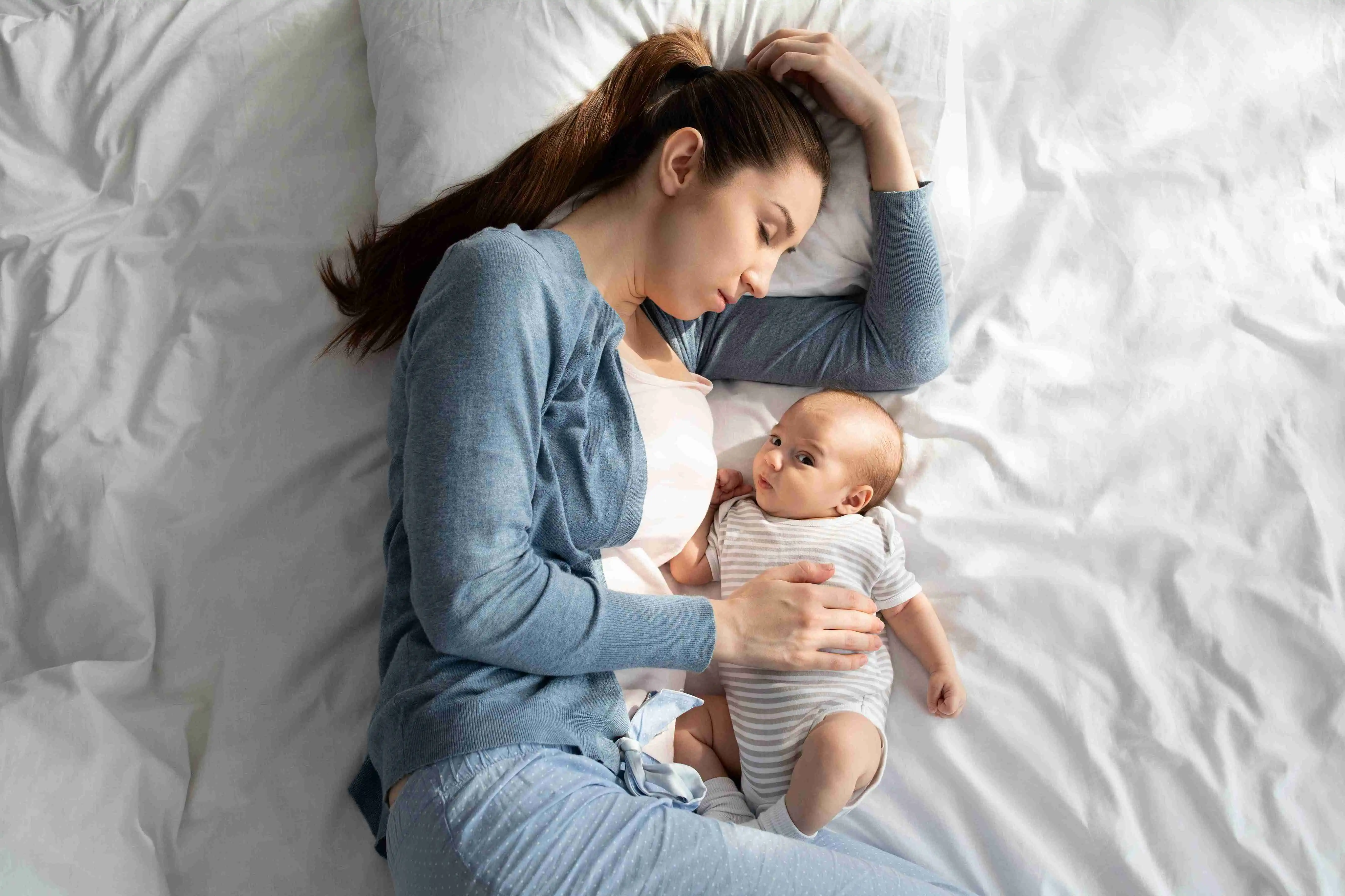 Tired Young Mother Sleeping in Bed While Her Adorable Newborn Child Awake