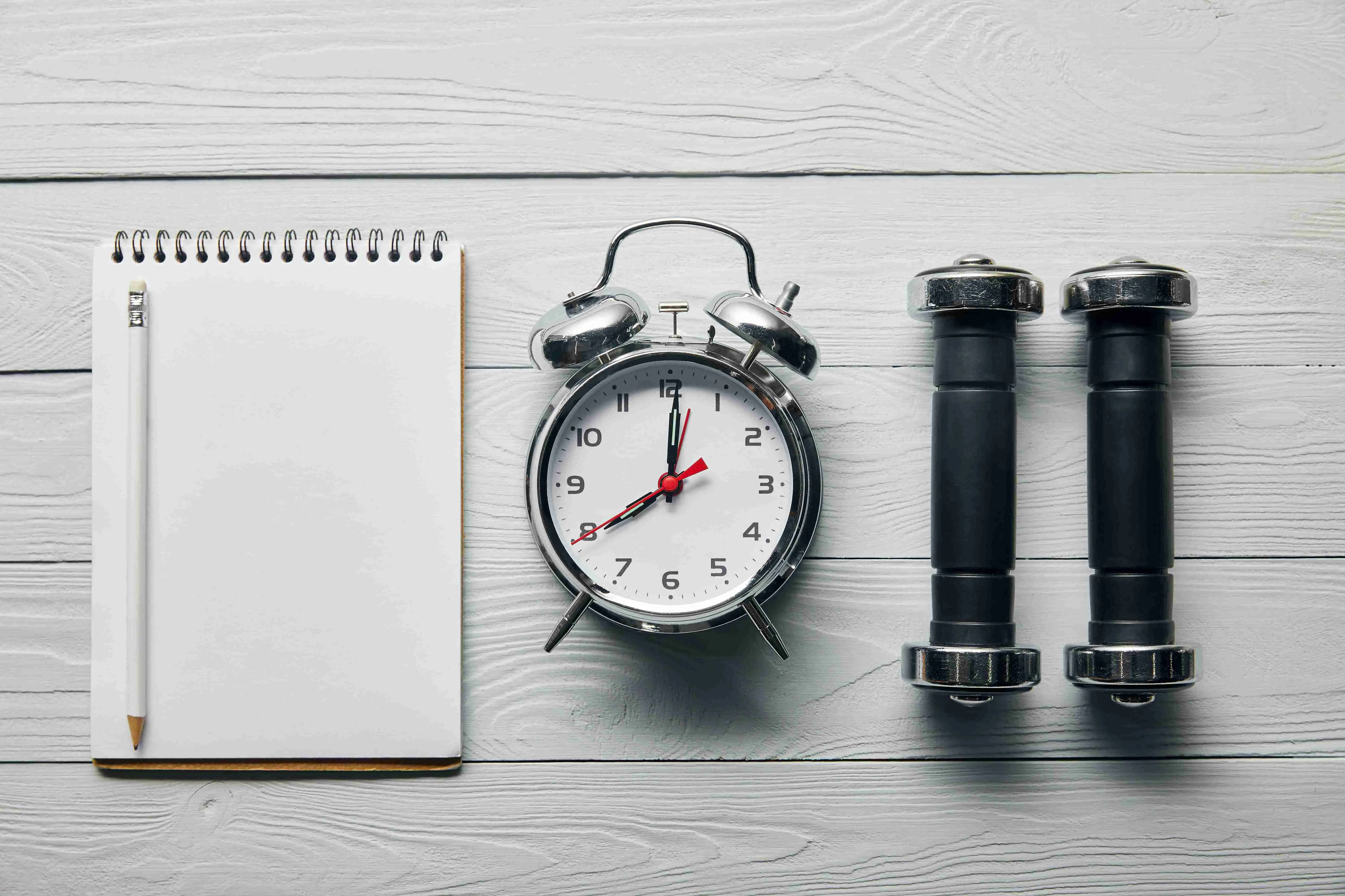 flat lay with silver alarm clock, empty notebook with pencil and dumbbells on wooden white