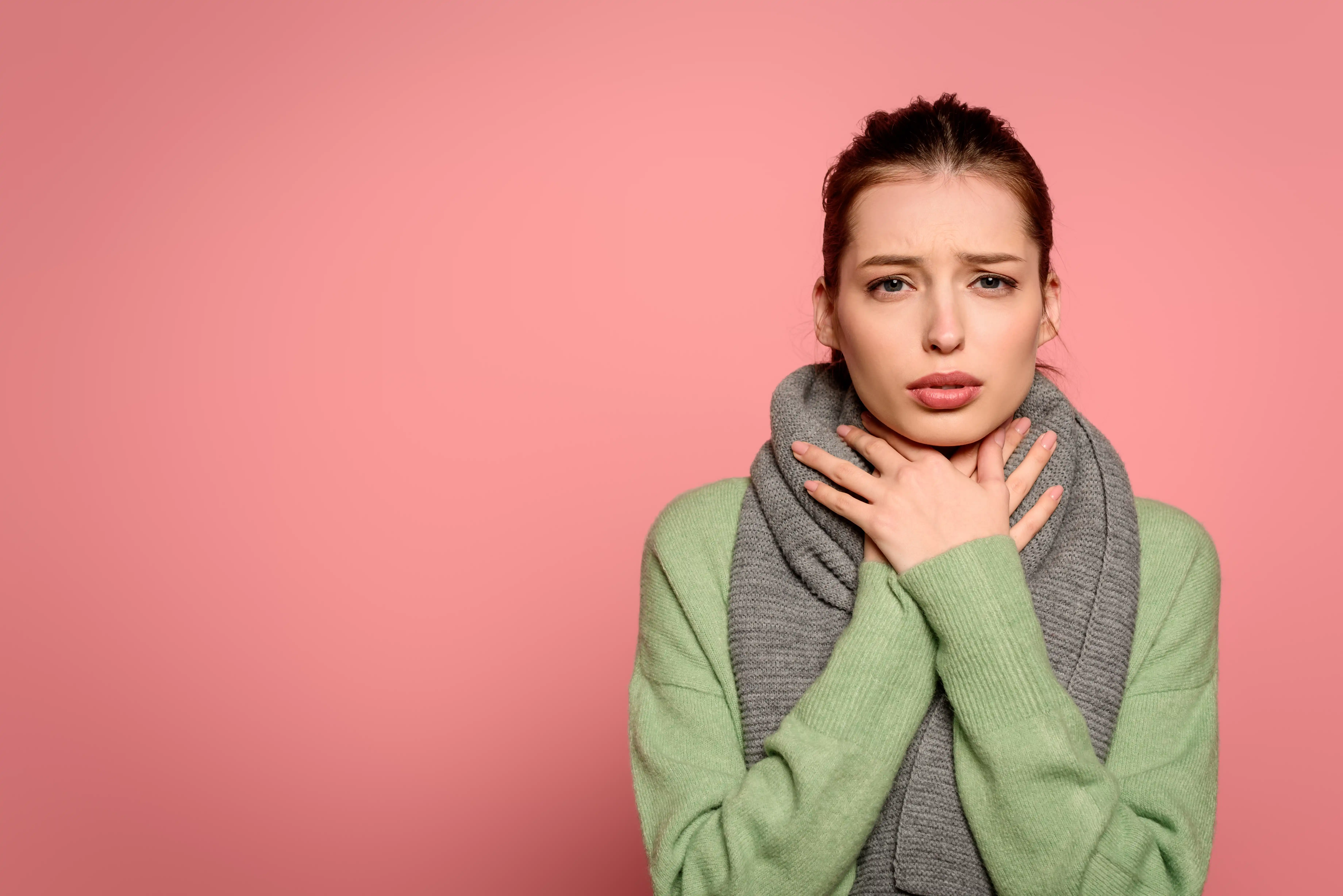 sick girl with sore throat touching neck wrapped in warm scarf isolated on pink