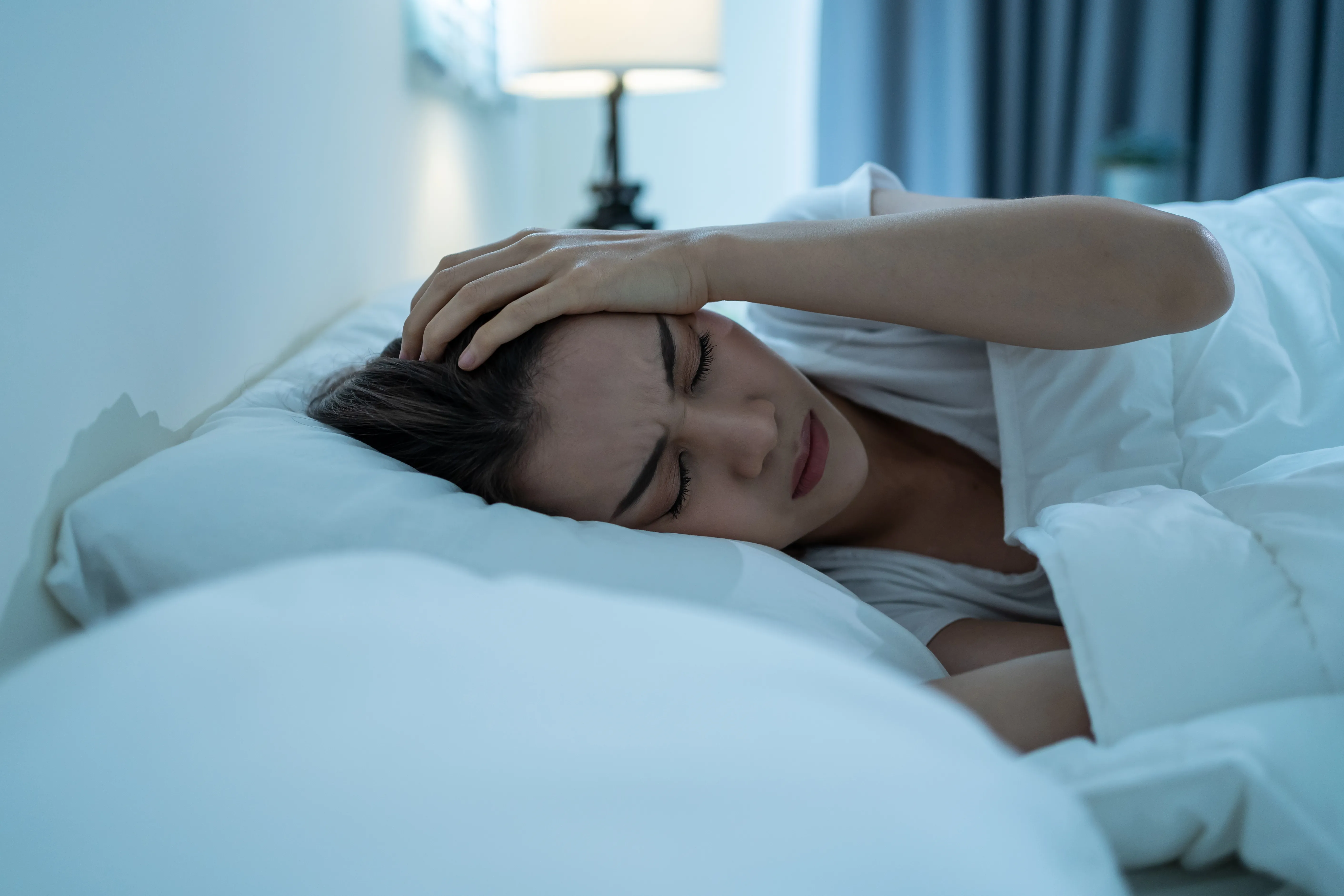 Woman lying in bed at night holding her head with a pained expression, eyes closed tightly, appearing to suffer from dizziness or vertigo while trying to sleep.