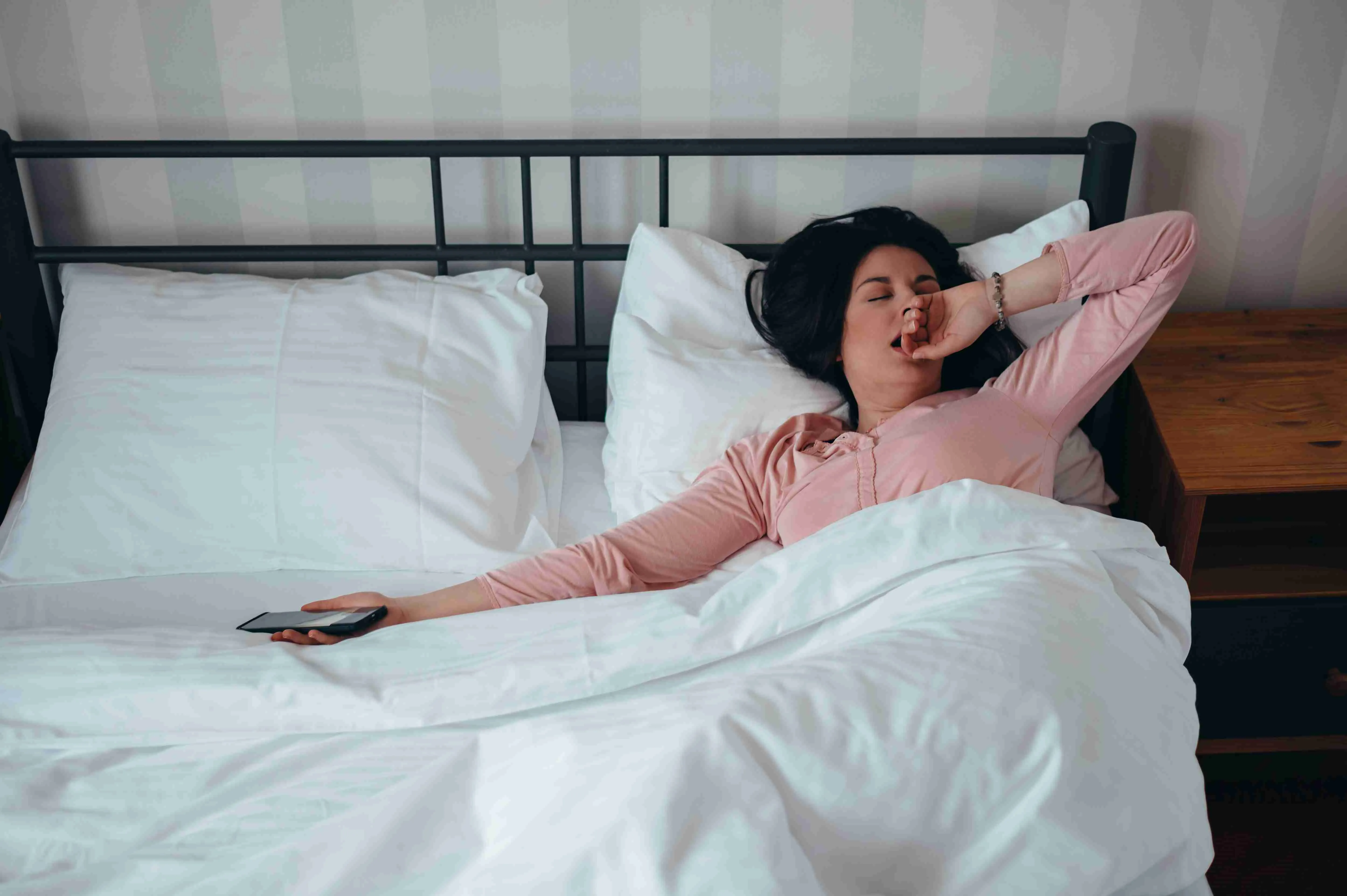 Woman waking up in her bed at home while holding a smartphone in her hand