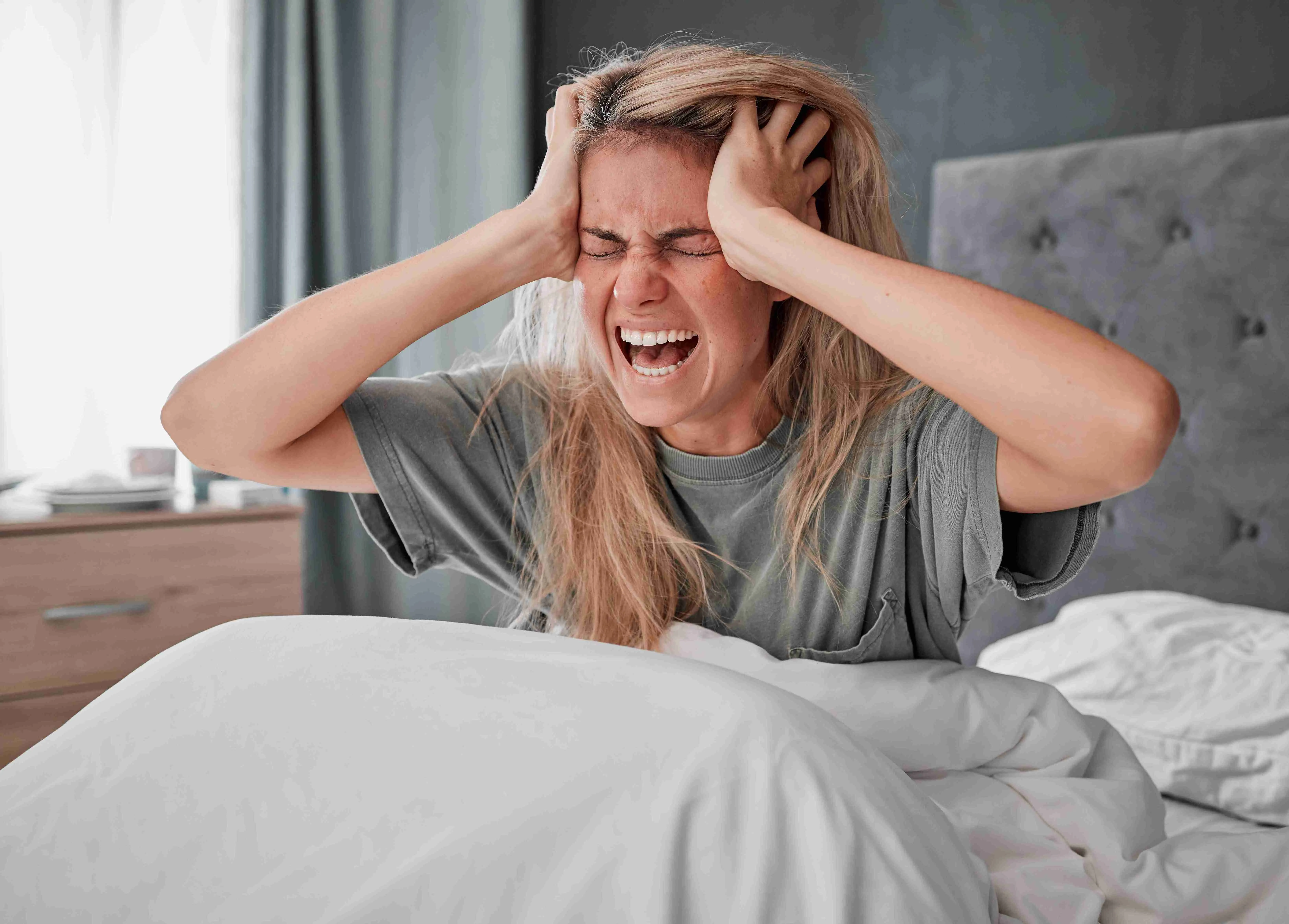 Person sitting on bed, holding their head, screaming, showing signs of a panic attack or extreme anxiety.