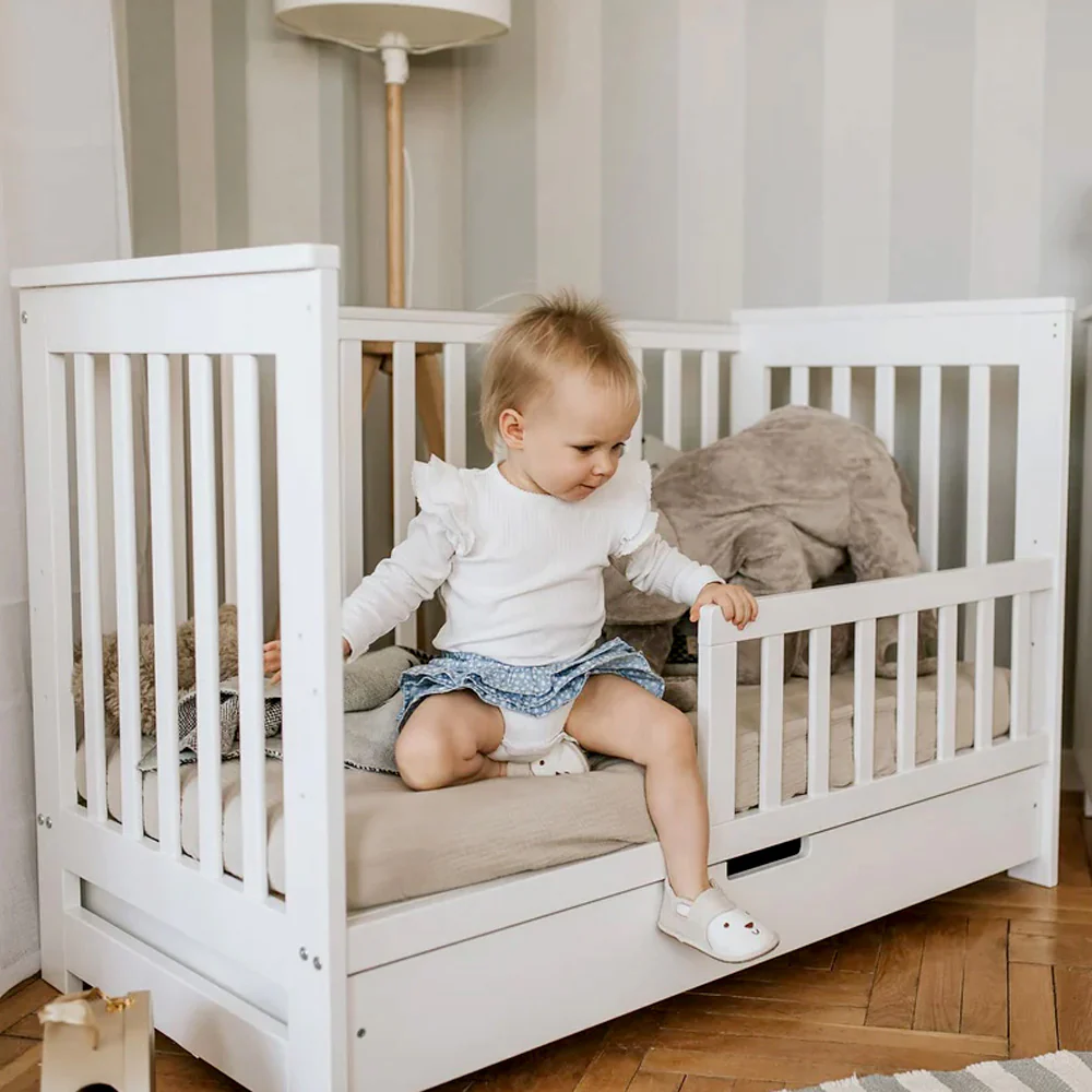 A toddler is sitting in a white wooden crib in a cozy room.