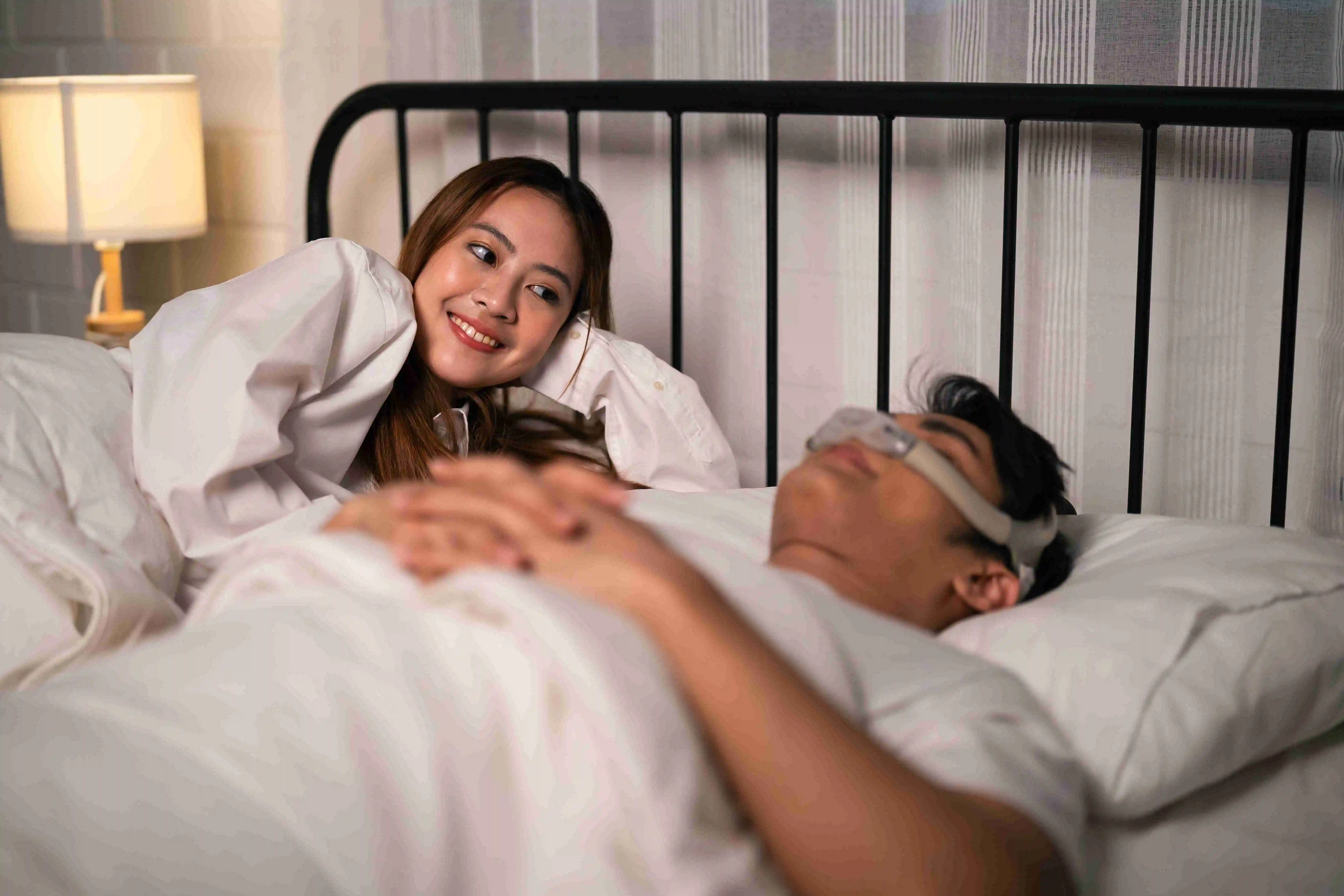 Happy Asian wife looking at husband wearing cpap machine while sleeping.