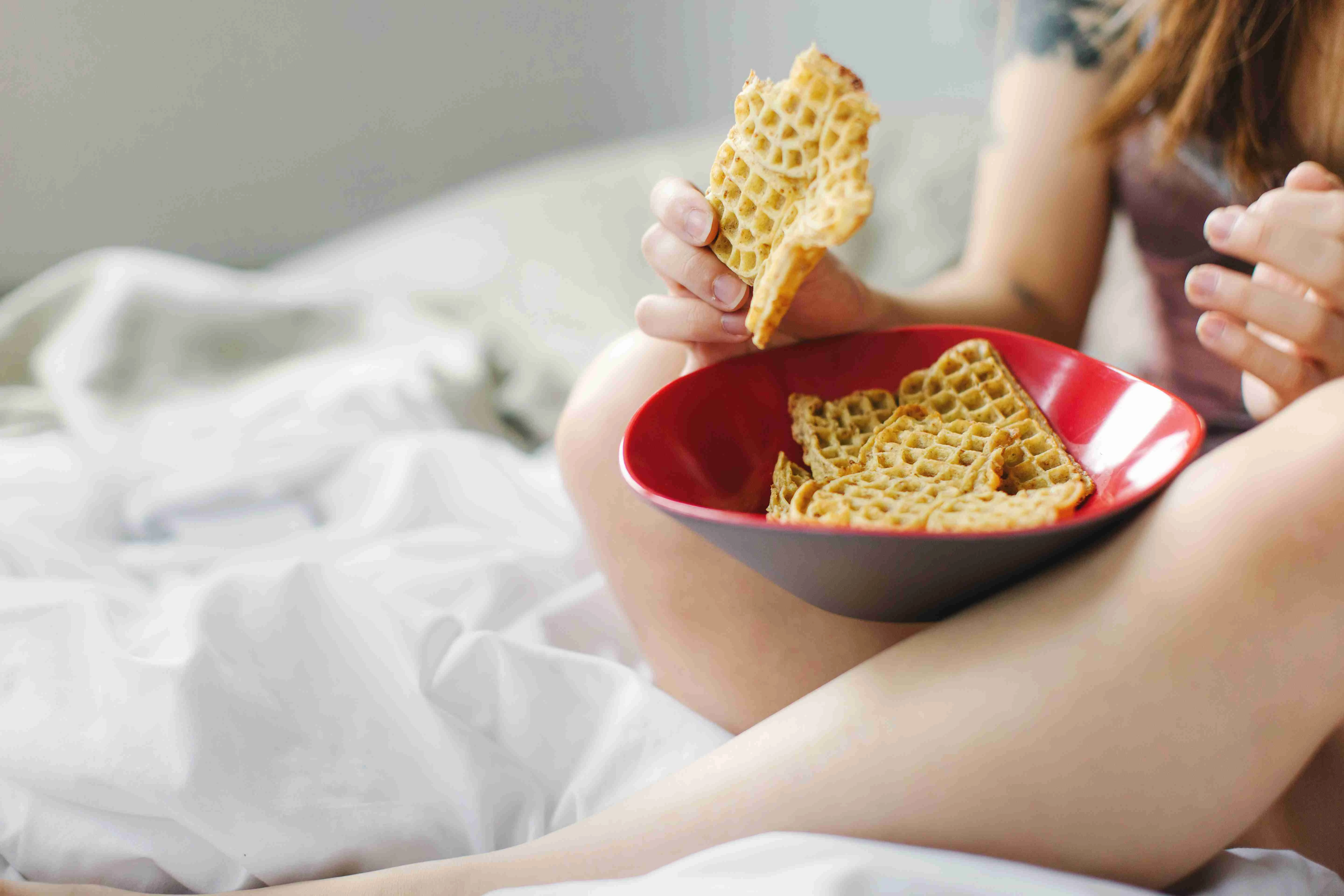 Young woman, sitting on bed, eating snack food from bowl