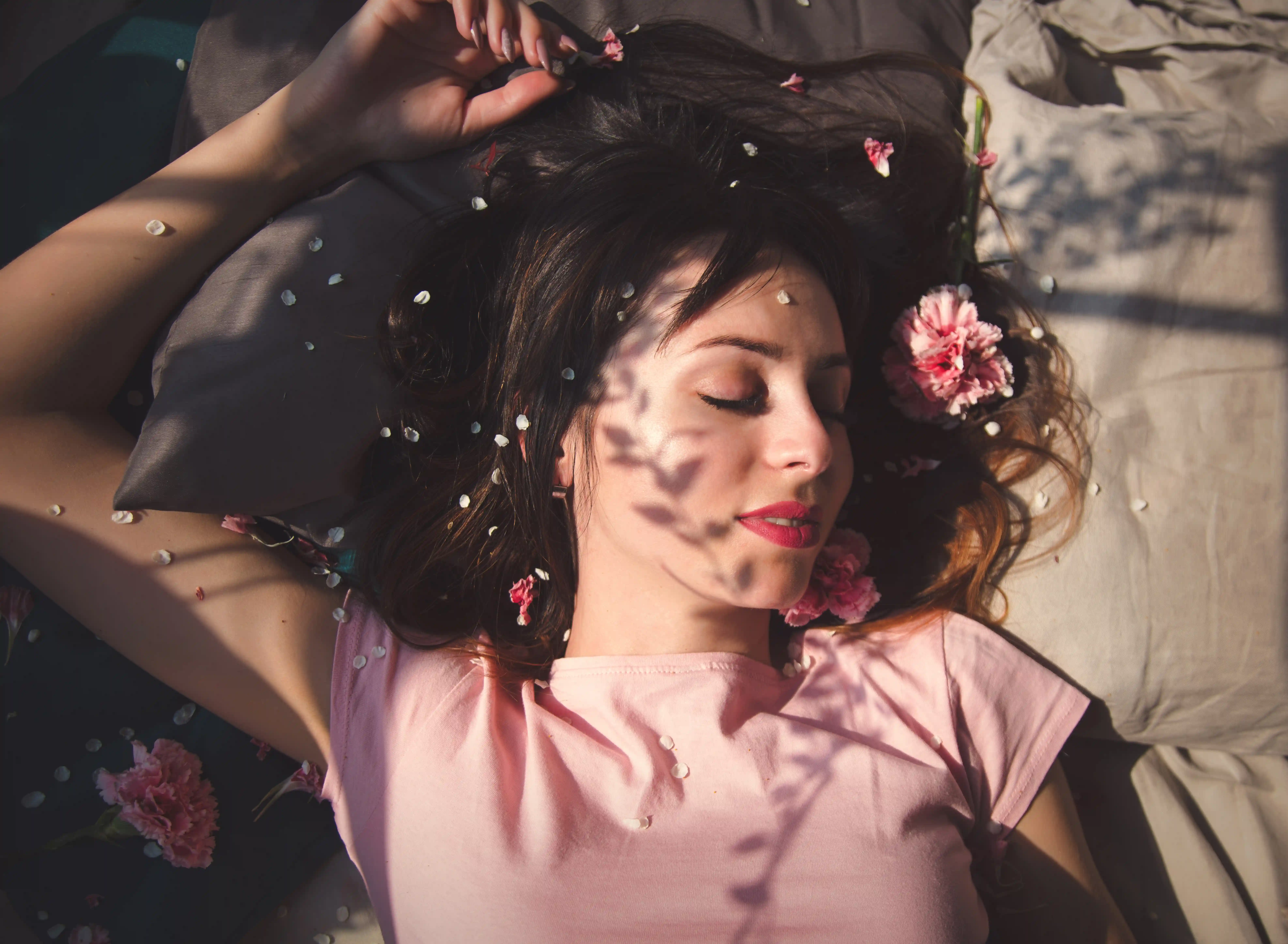 Brunette woman asleep among flower petals with shadows across her face.