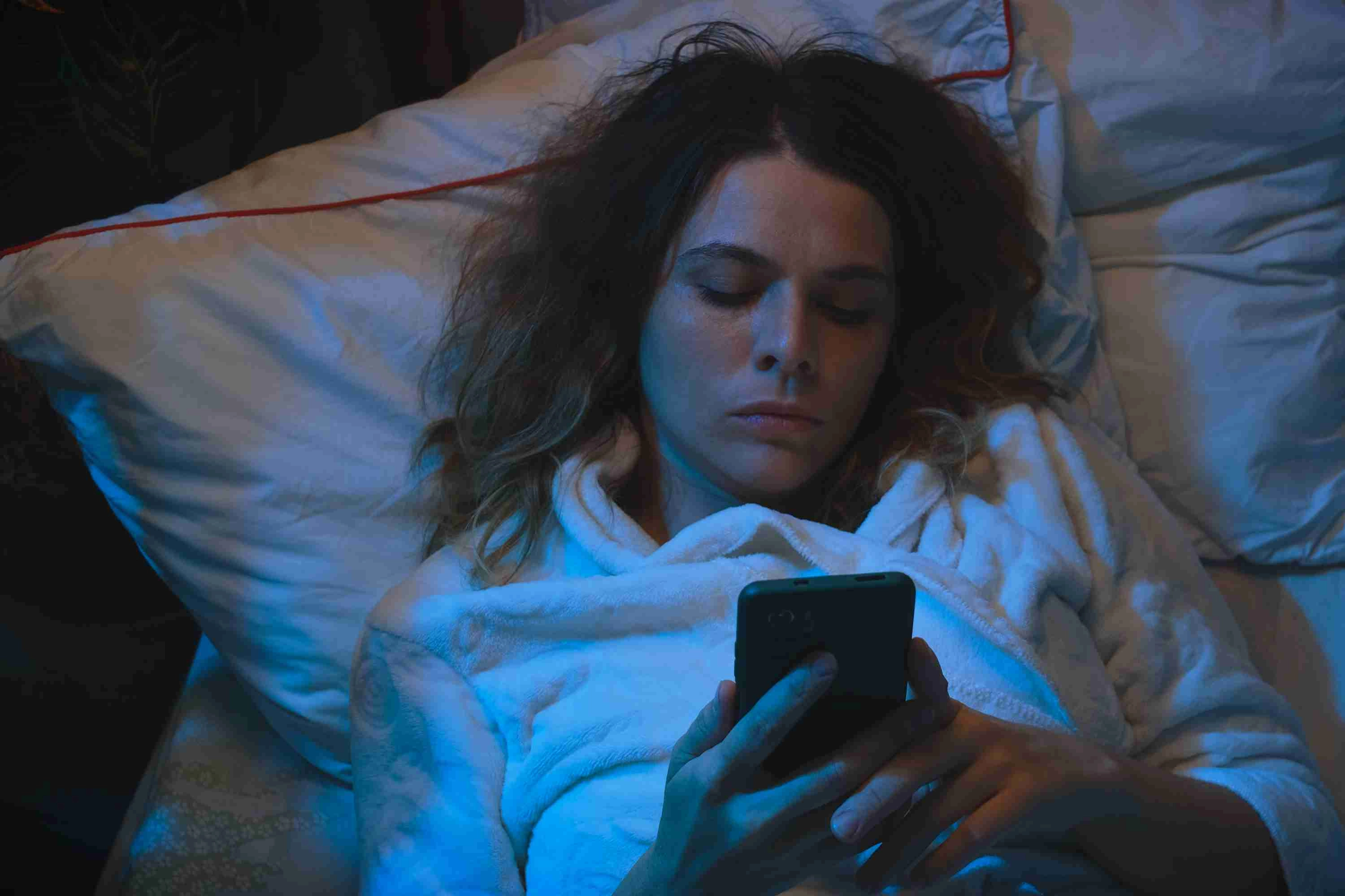 Young woman cannot fall asleep, using smartphone lying in bed at night.