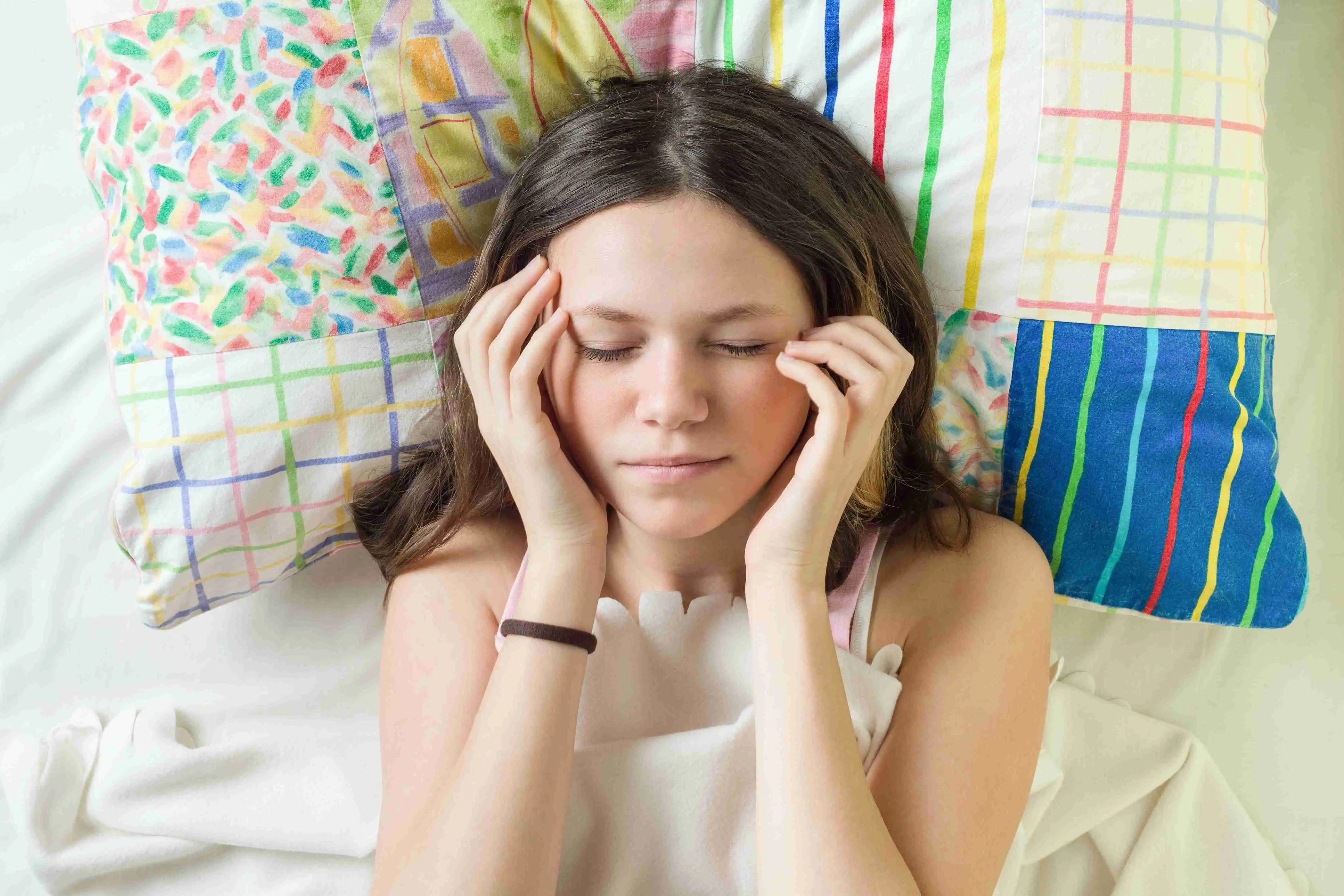 Close-up face of teenage girl on pillow with closed eyes holding head with hands