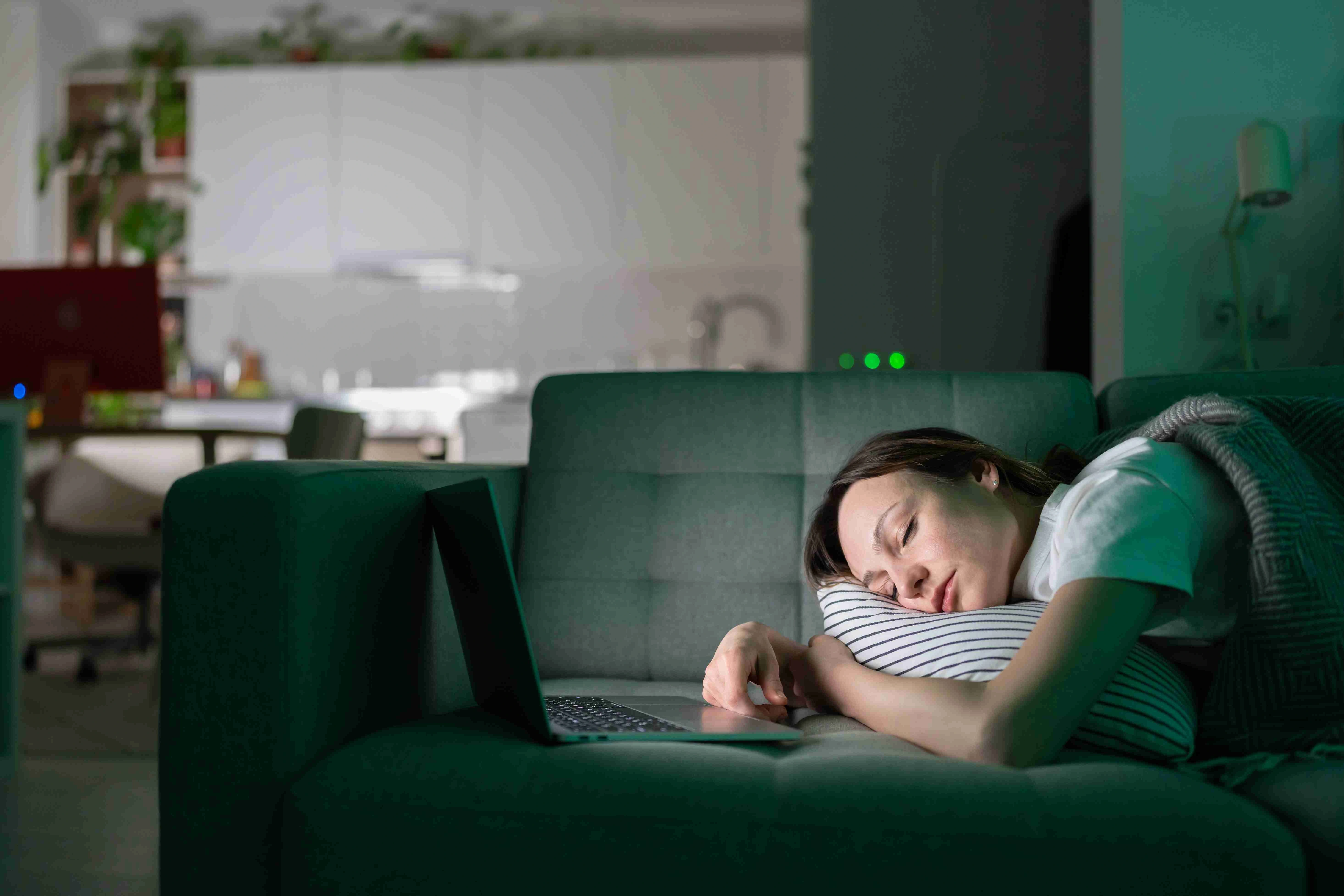 Exhausted young woman sleeping on couch after working with laptop computer late at night at home