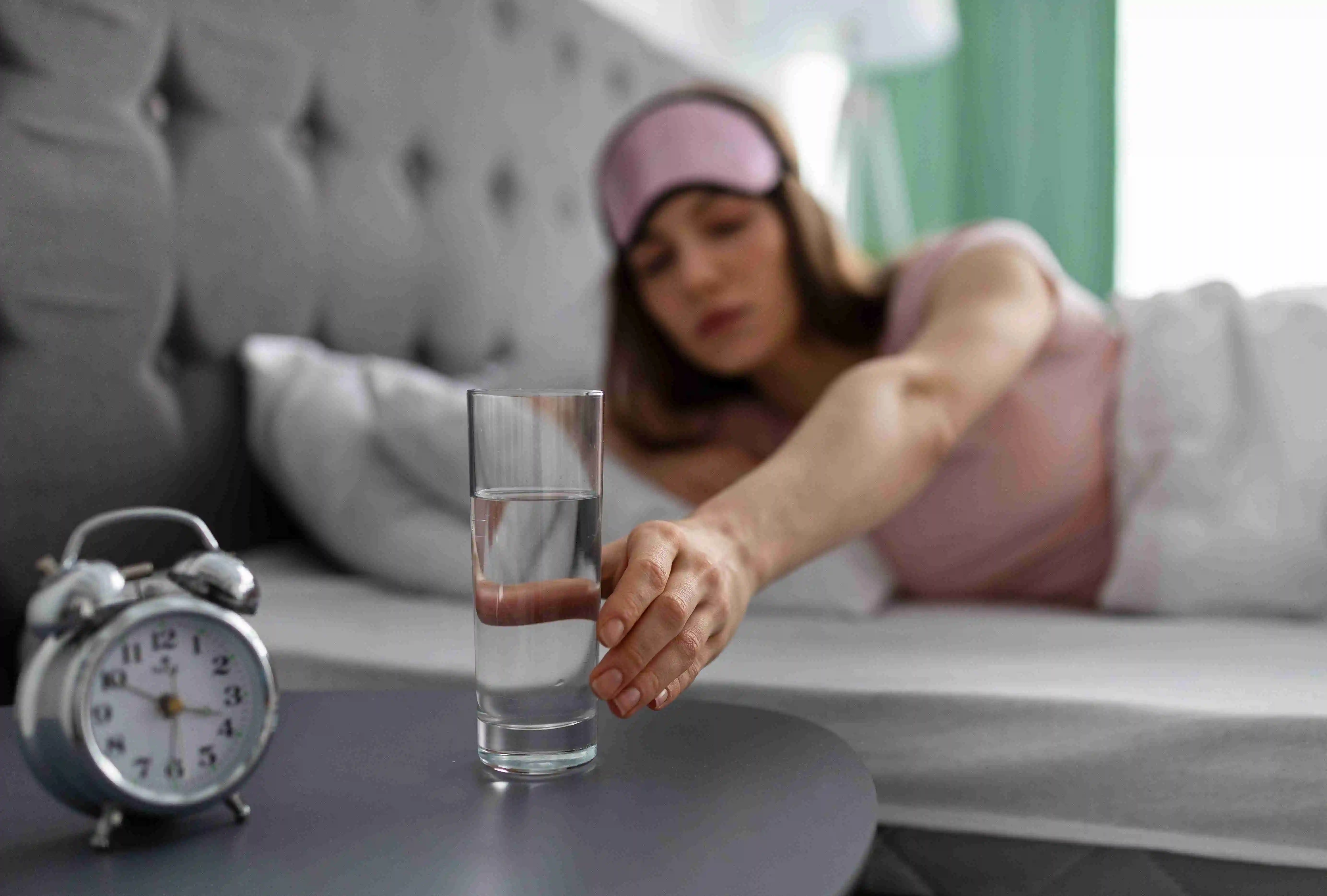 Young woman in sleep mask taking glass of mineral water from bedside table while lying in bed at