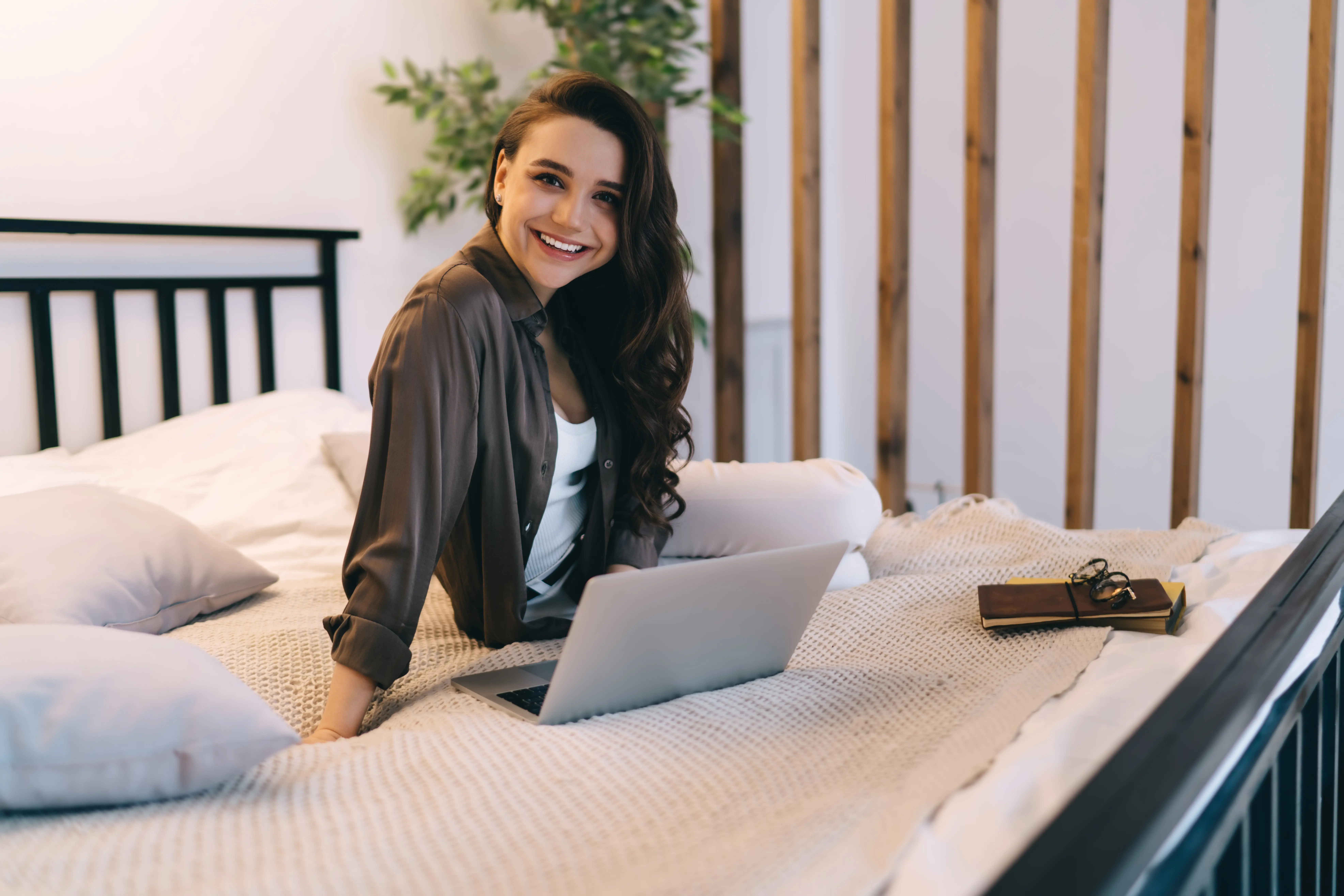 Positive young female student relaxing on comfortable bed with smartphone while looking at camera and smiling