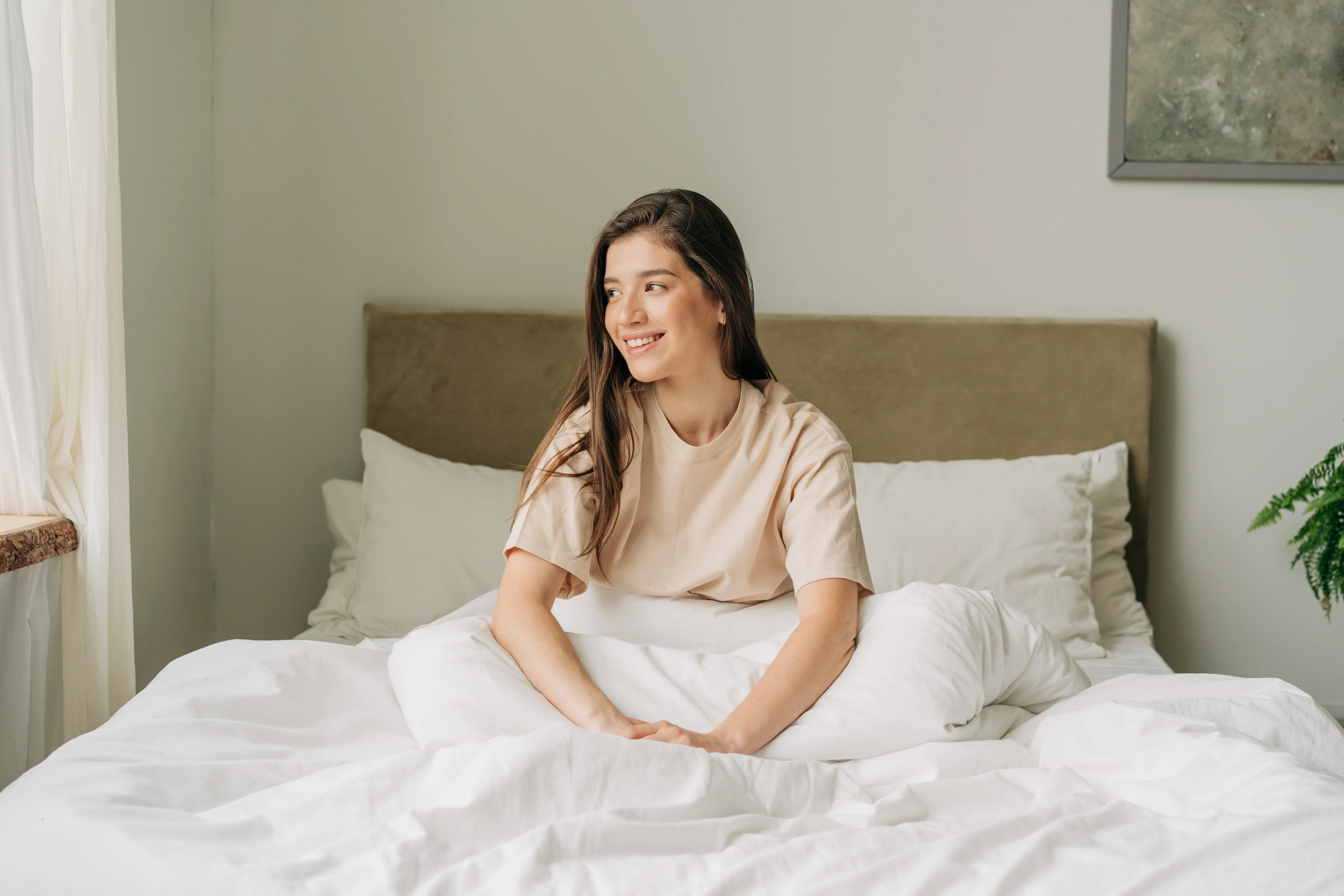 Young smiling rested brunette woman sitting on the bed in a cozy bright bedroom early in the morning