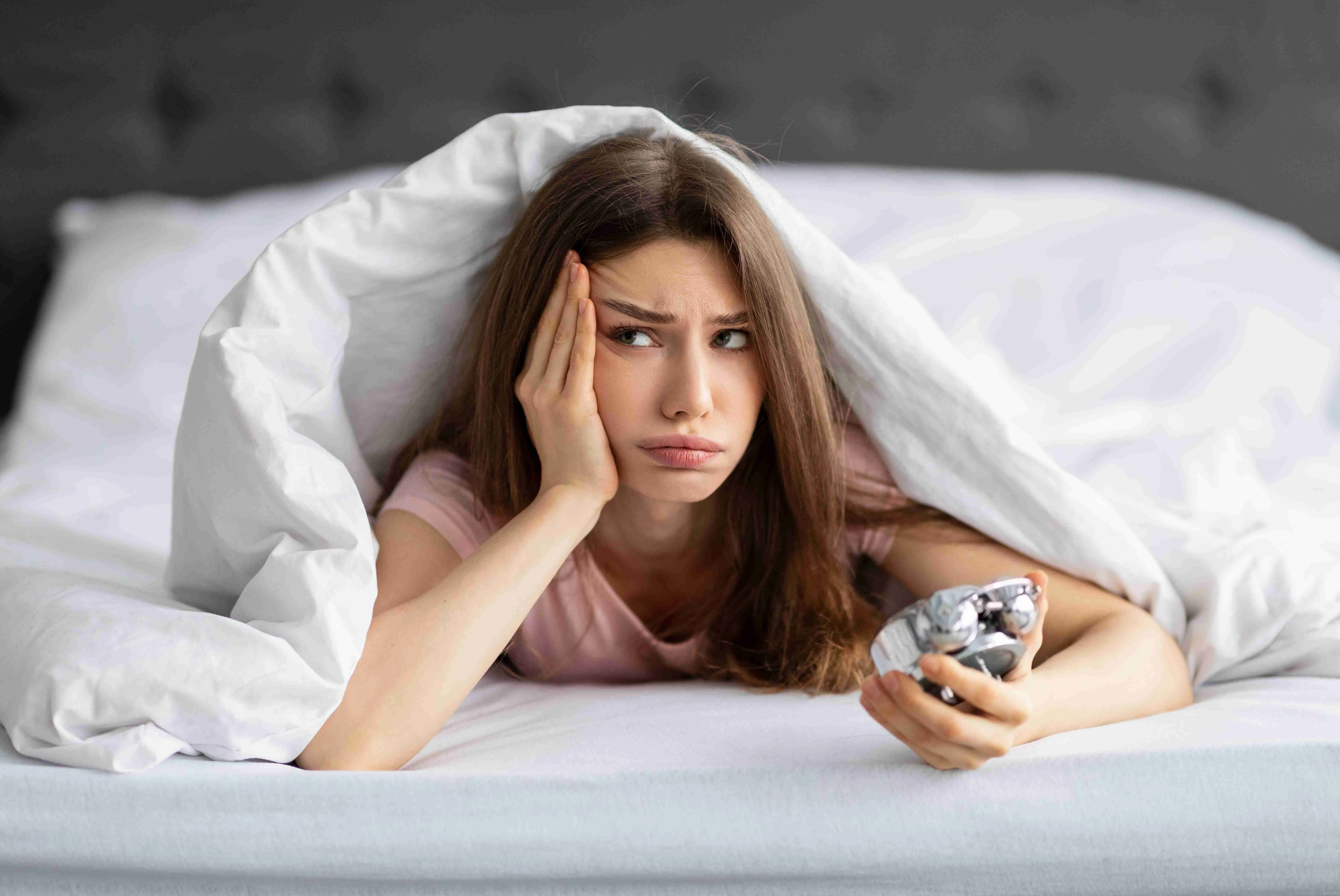 Unhappy young Caucasian woman lying in bed with alarm clock