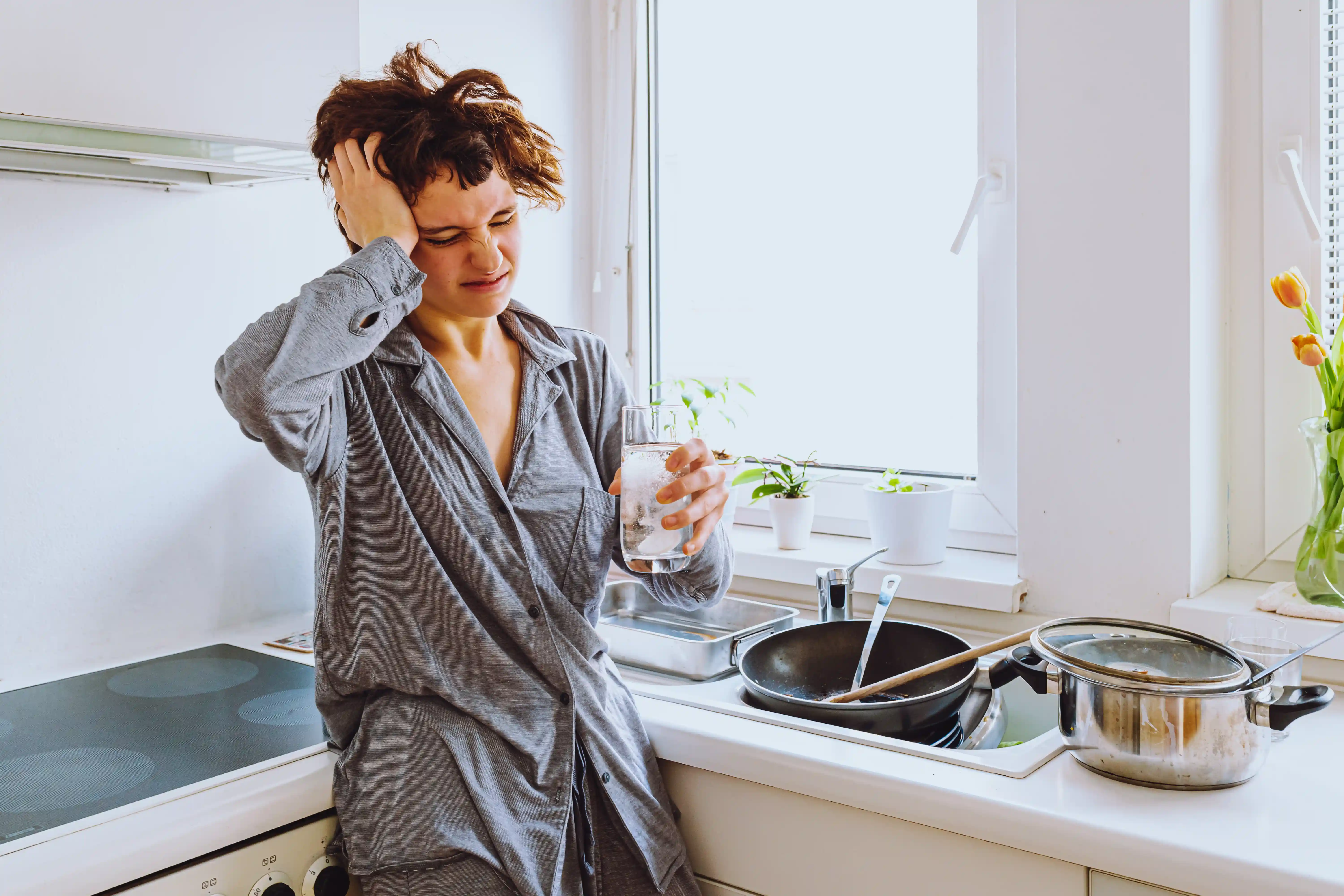 teenage girl in pajamas, with disheveled hair, with headache, fatigue, depression, stands in kitchen, holds glass of water and aspirin tablet in hands, against blurred background of unwashed dishes, mess.