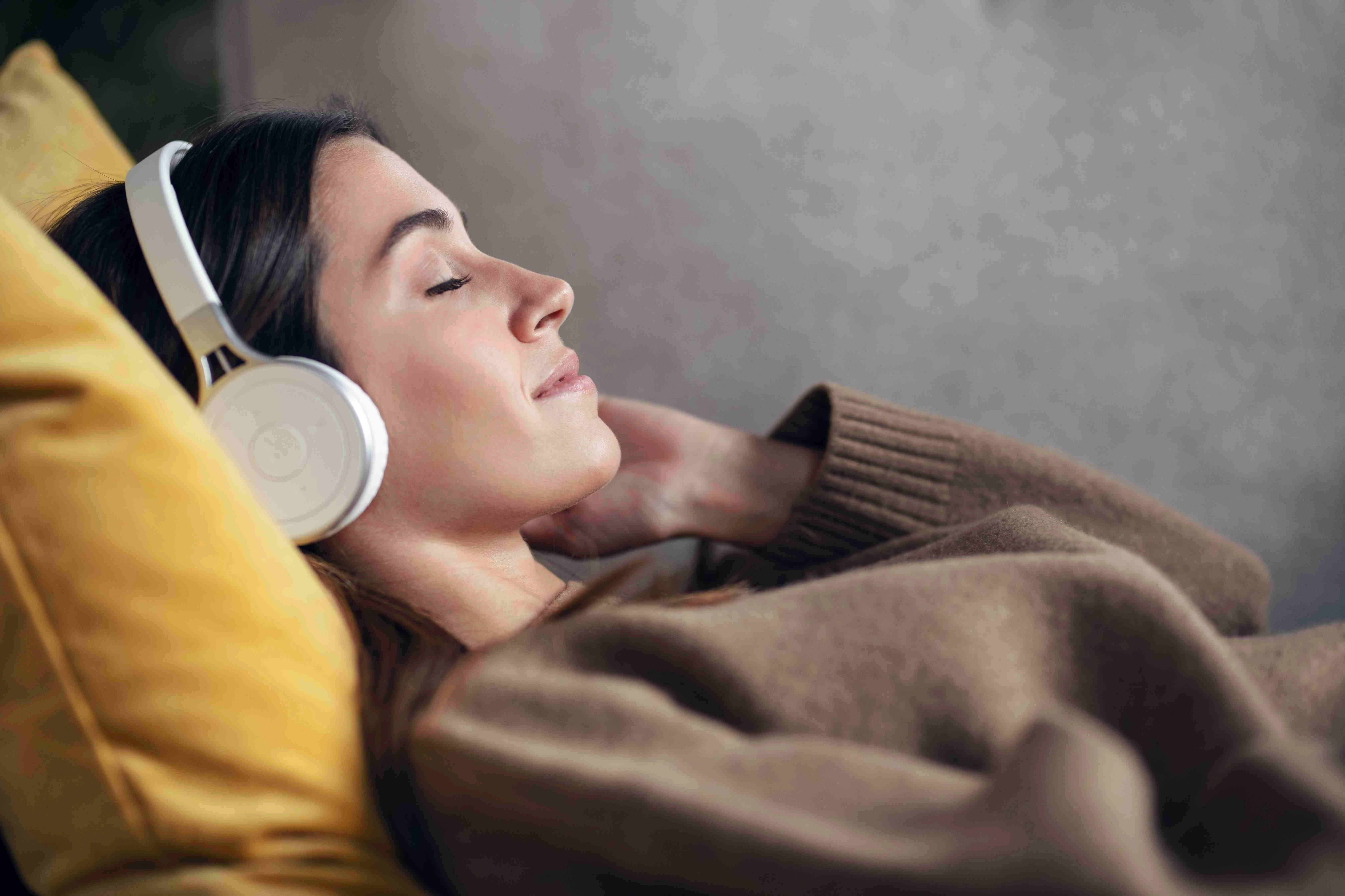 Beautiful woman listening to music with headphone while using smartphone lying on sofa at home.