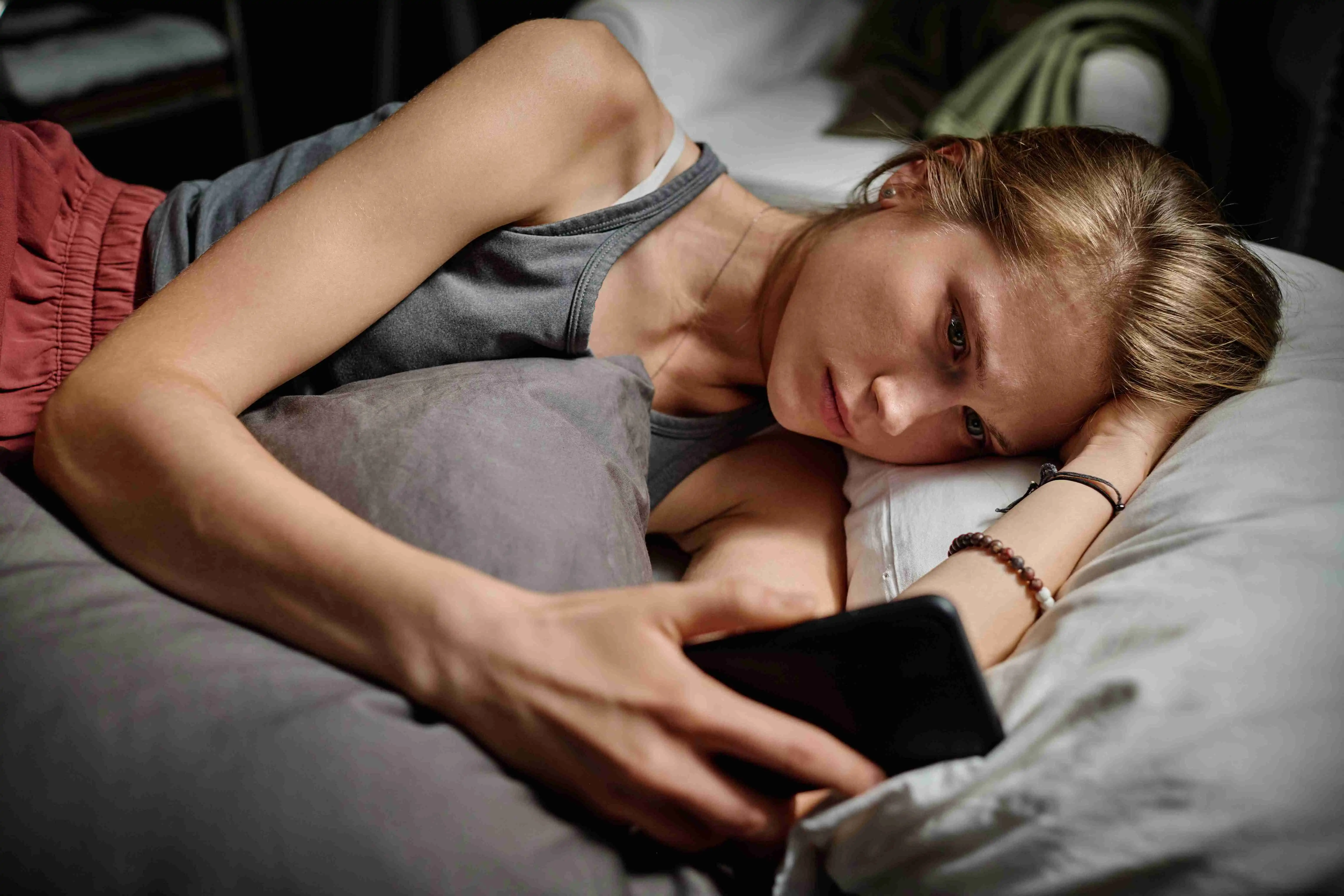 Depressed blond teenage girl looking through cyberbullying messages while lying in bed late at night and suffering from insomnia