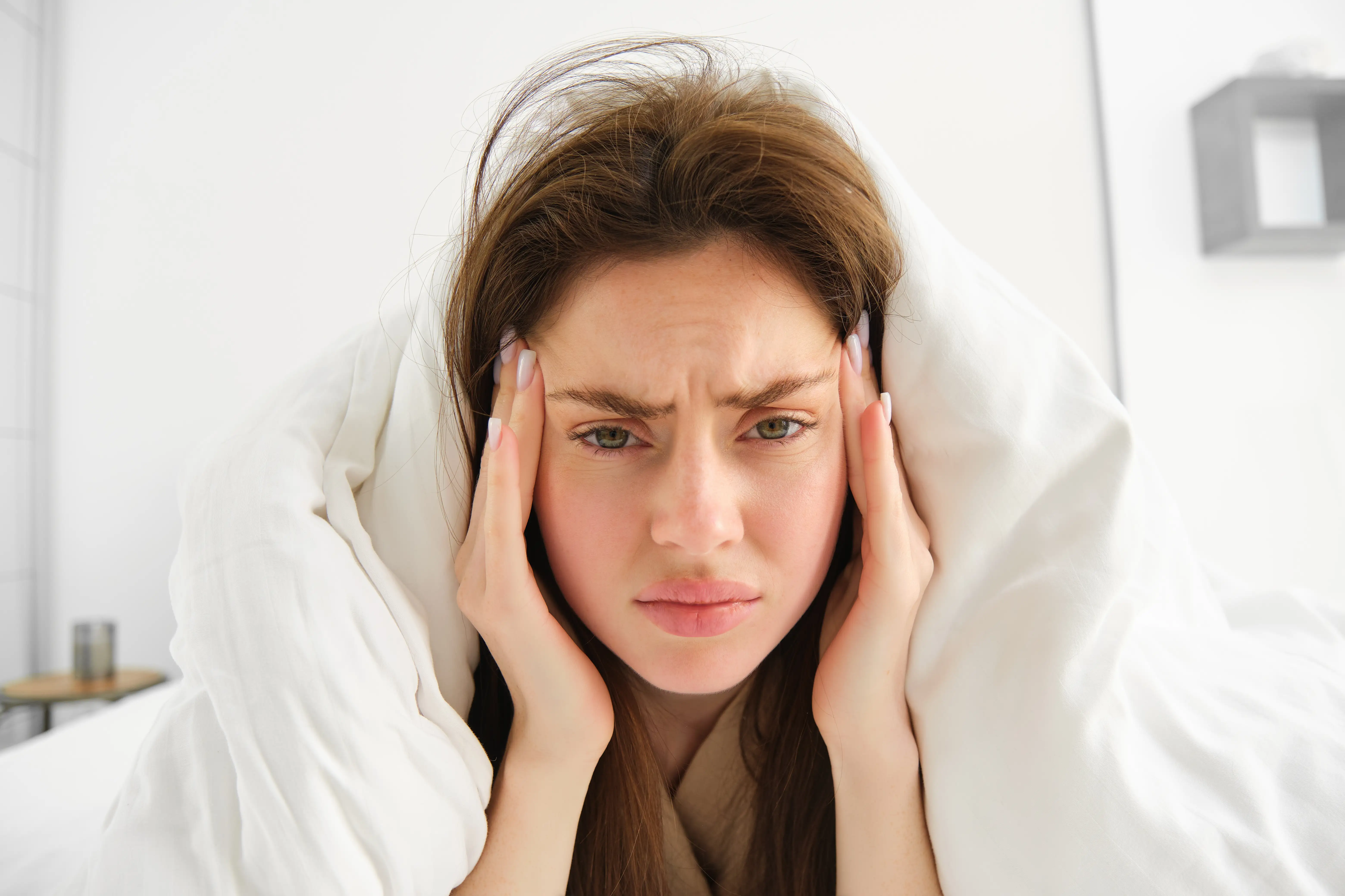 Woman feeling unwell in bed, lying in her bedroom under blanket and white sheets in morning