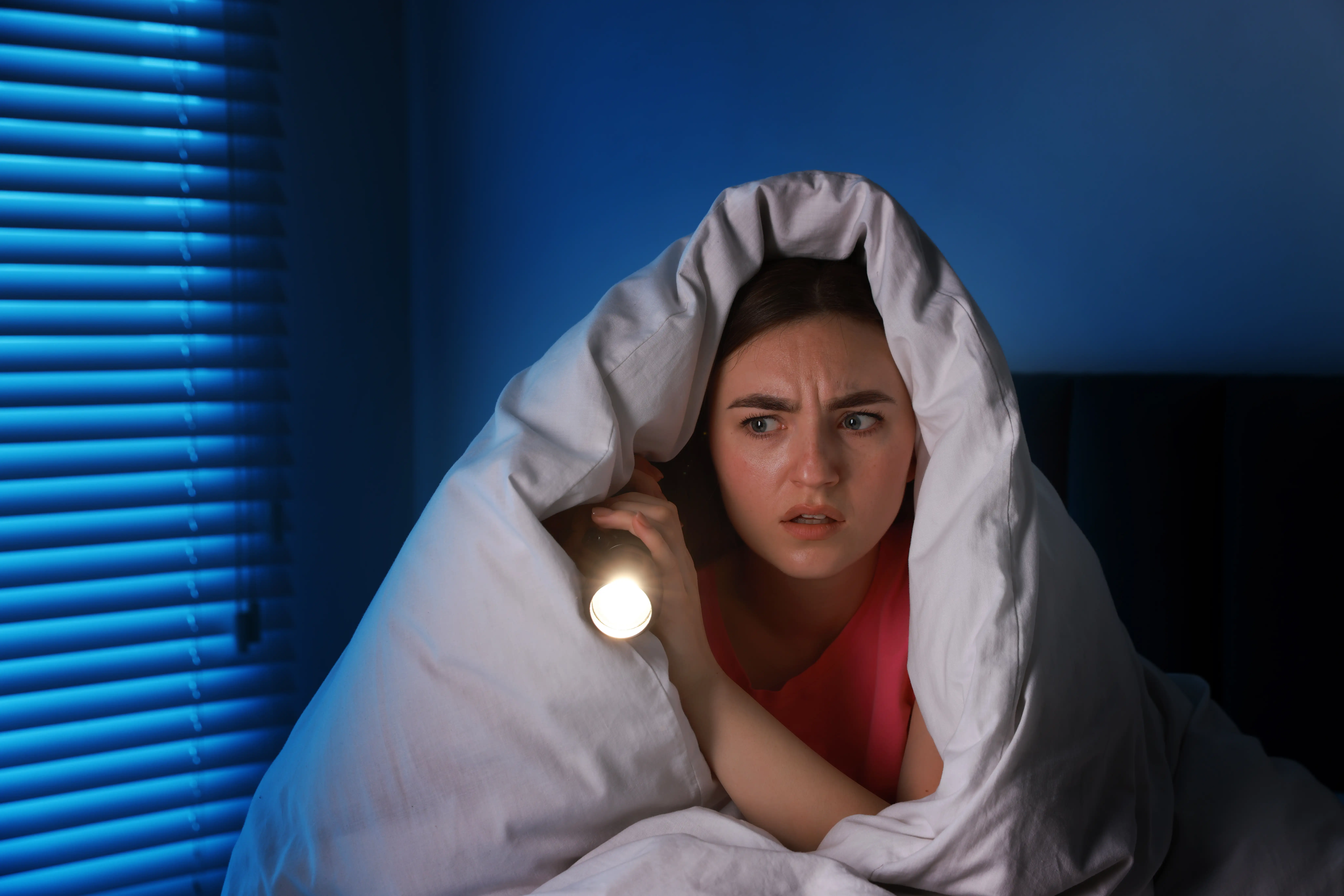 Fear of darkness. Scared young with blanket and flashlight on bed indoors at night