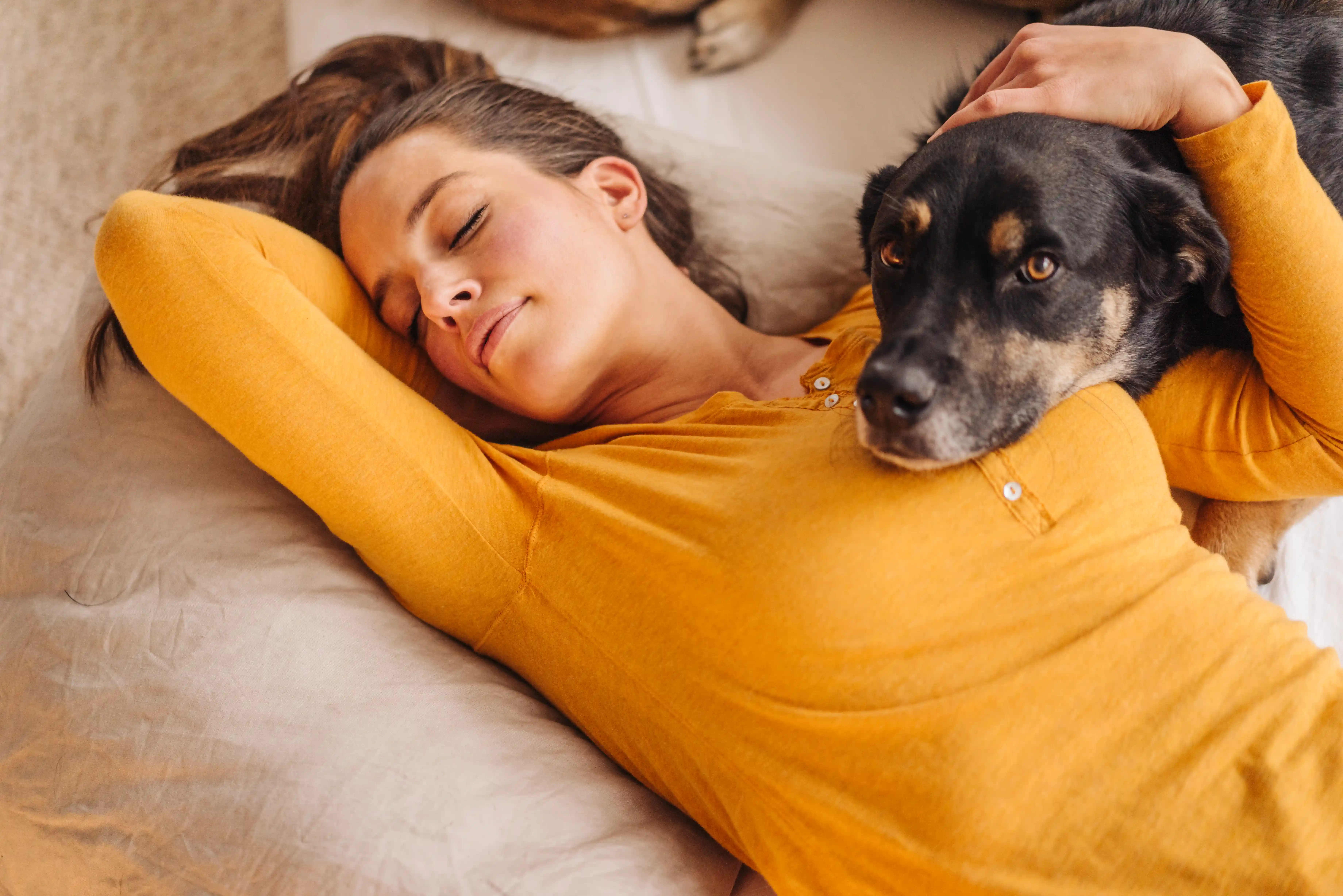 A woman with her light brown hair rests on a bed with a black and brown dog lying against her chest