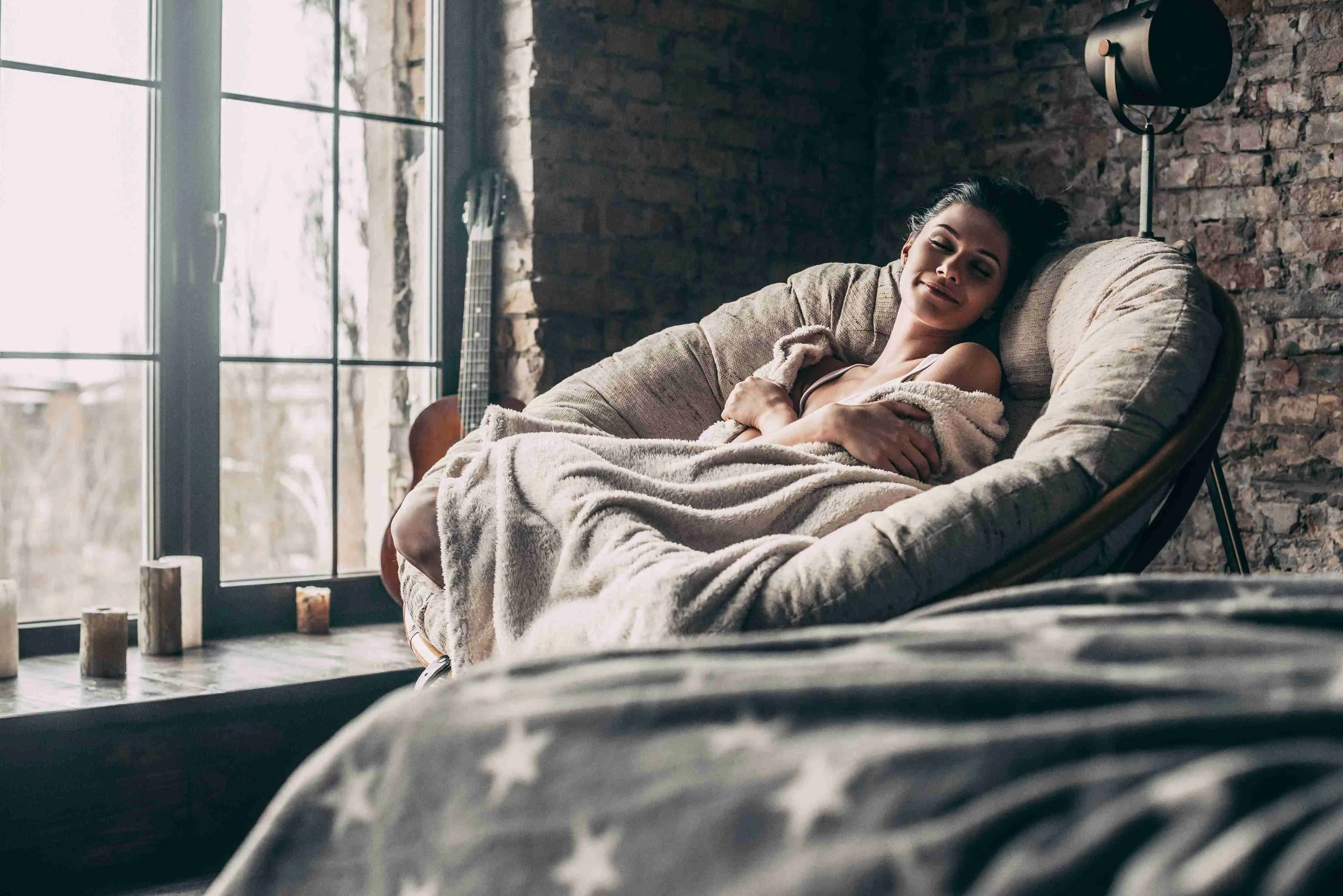 Attractive young woman covered with blanket sleeping and smiling while sitting in an armchair at home