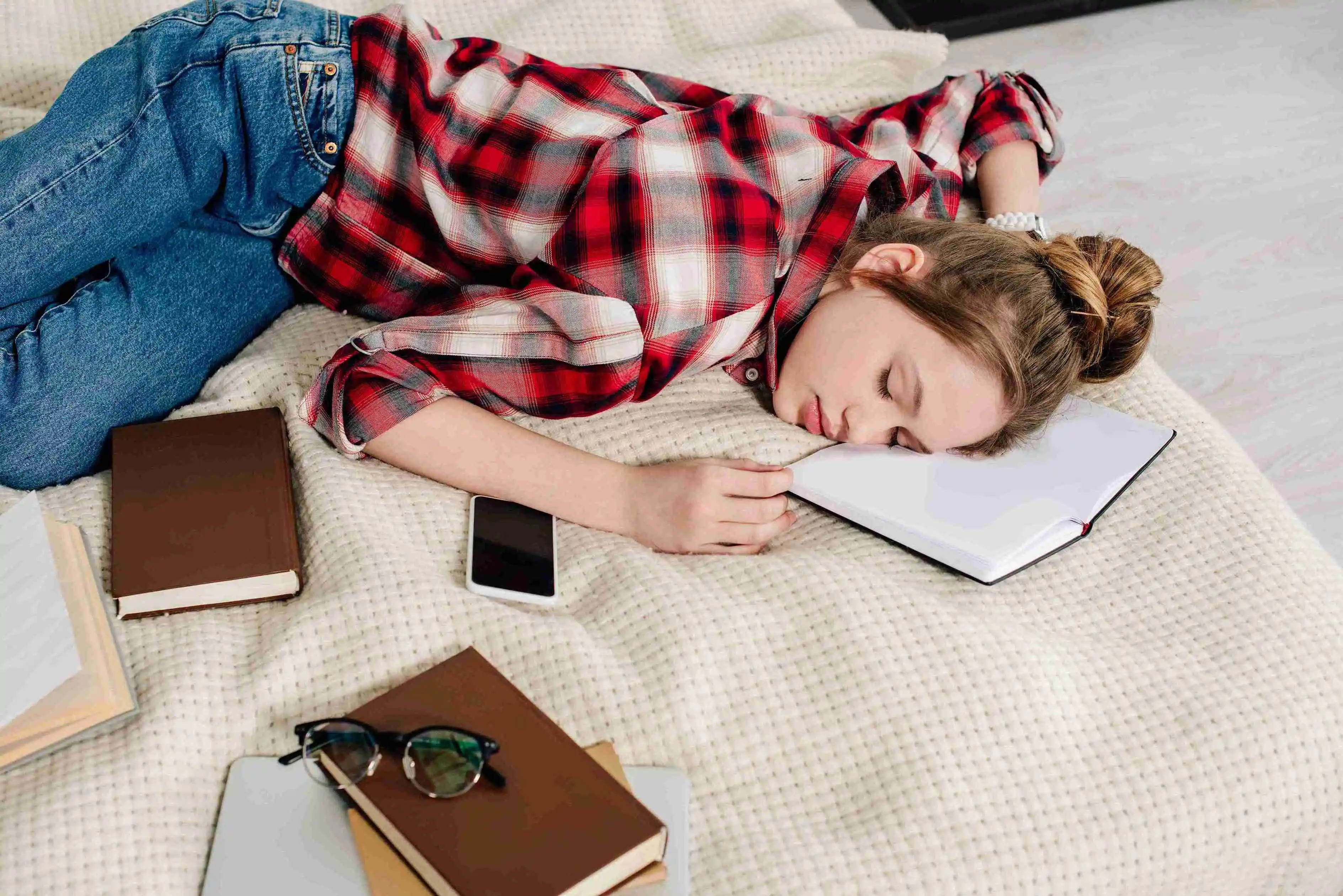 Teenager in checkered shirt sleeping on bed with books and smartphone
