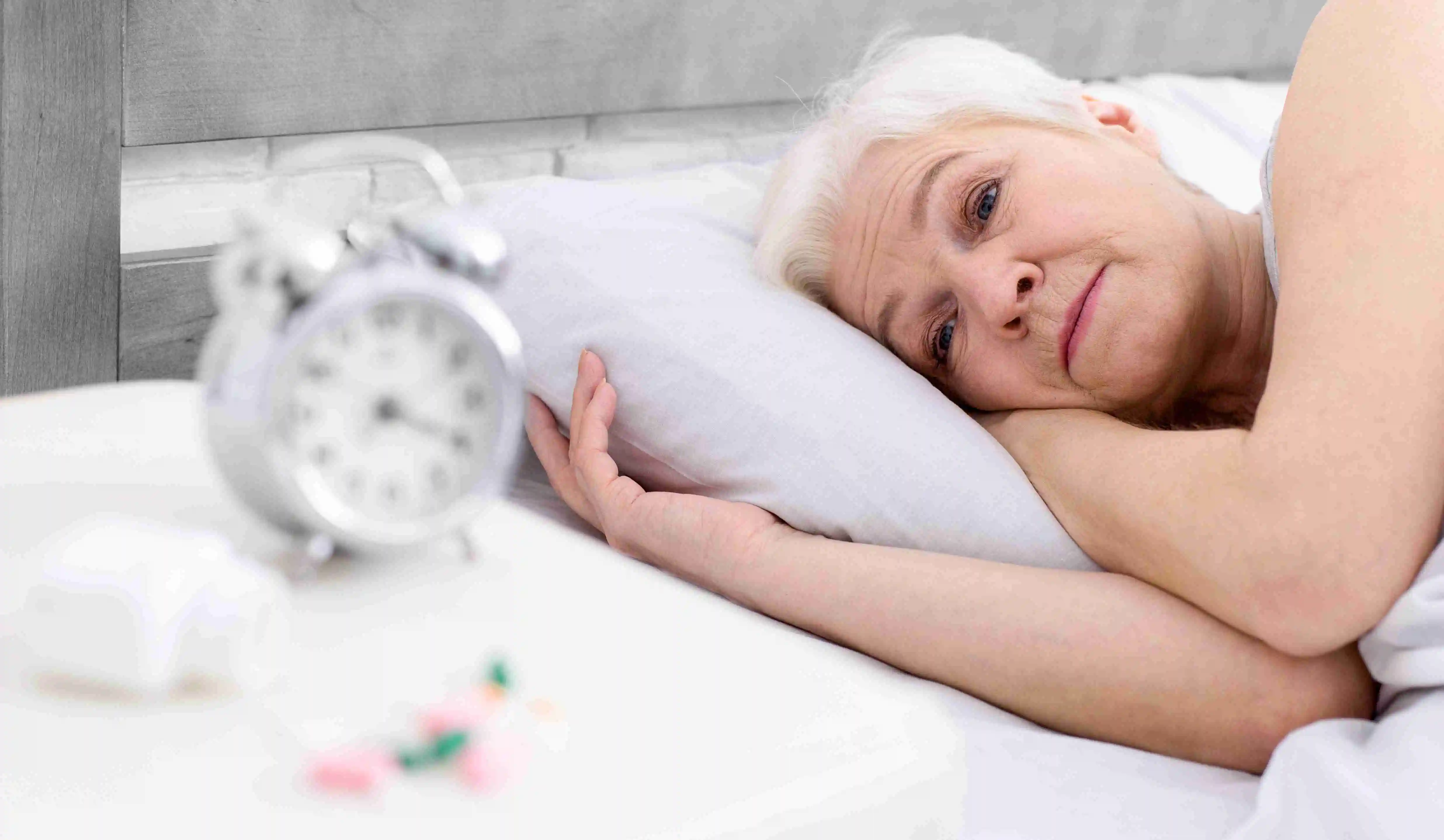 A woman is laying in bed, with her eyes open, next to an alarm clock set on the bedside table. The clock displays a time indicating the morning hour.