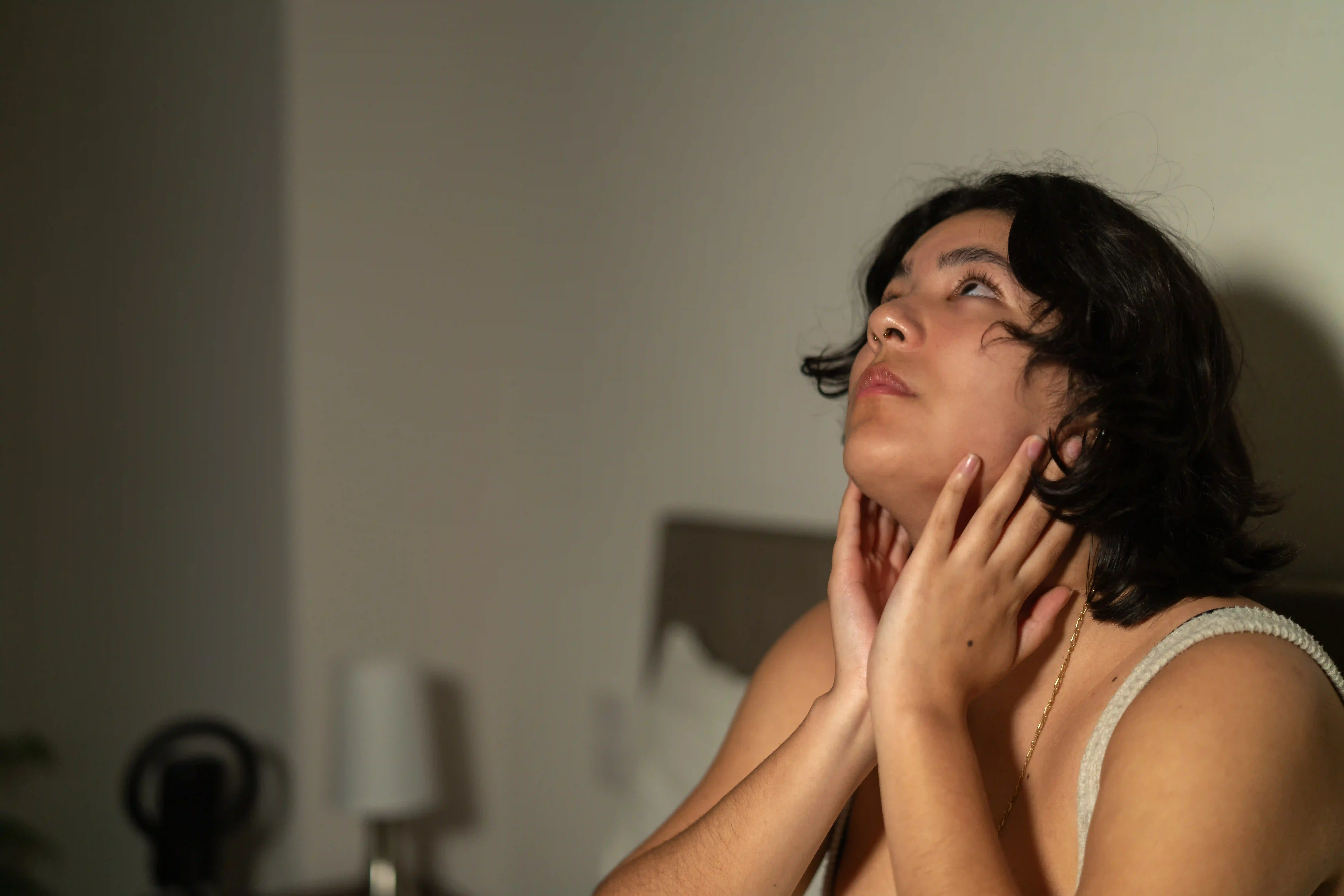 Young woman sitting on bed, suffering from insomnia and experiencing stress at night, touching her neck with hands