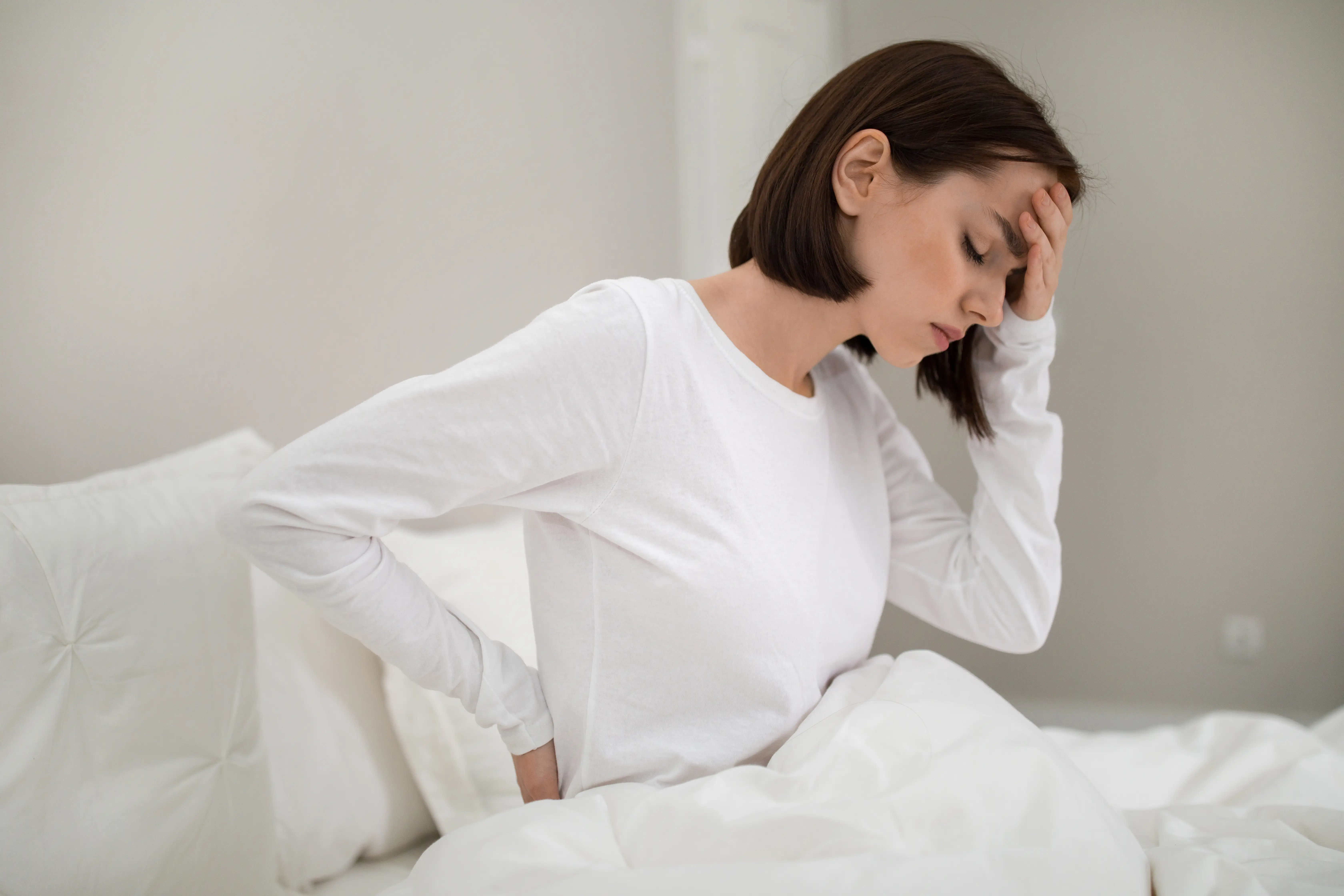 Tired youg lady suffering from back pain after sleeping in bedroom, waking up in morning, sitting on bed and touching her lower back and forehead, wearing white pajamas, copy space, side view