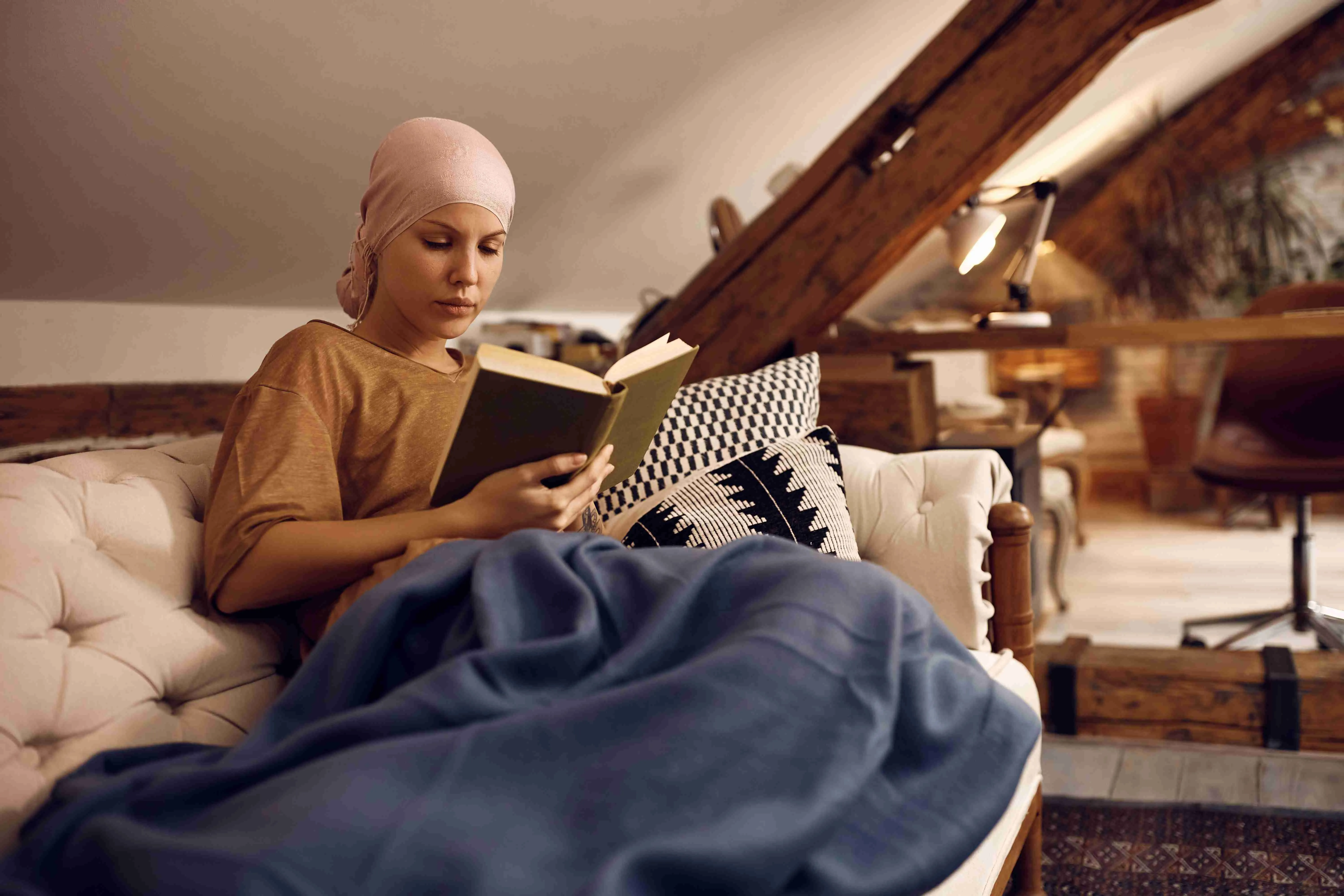 Young woman with cancer reading a novel and wearing headscarf while relaxing on the sofa.