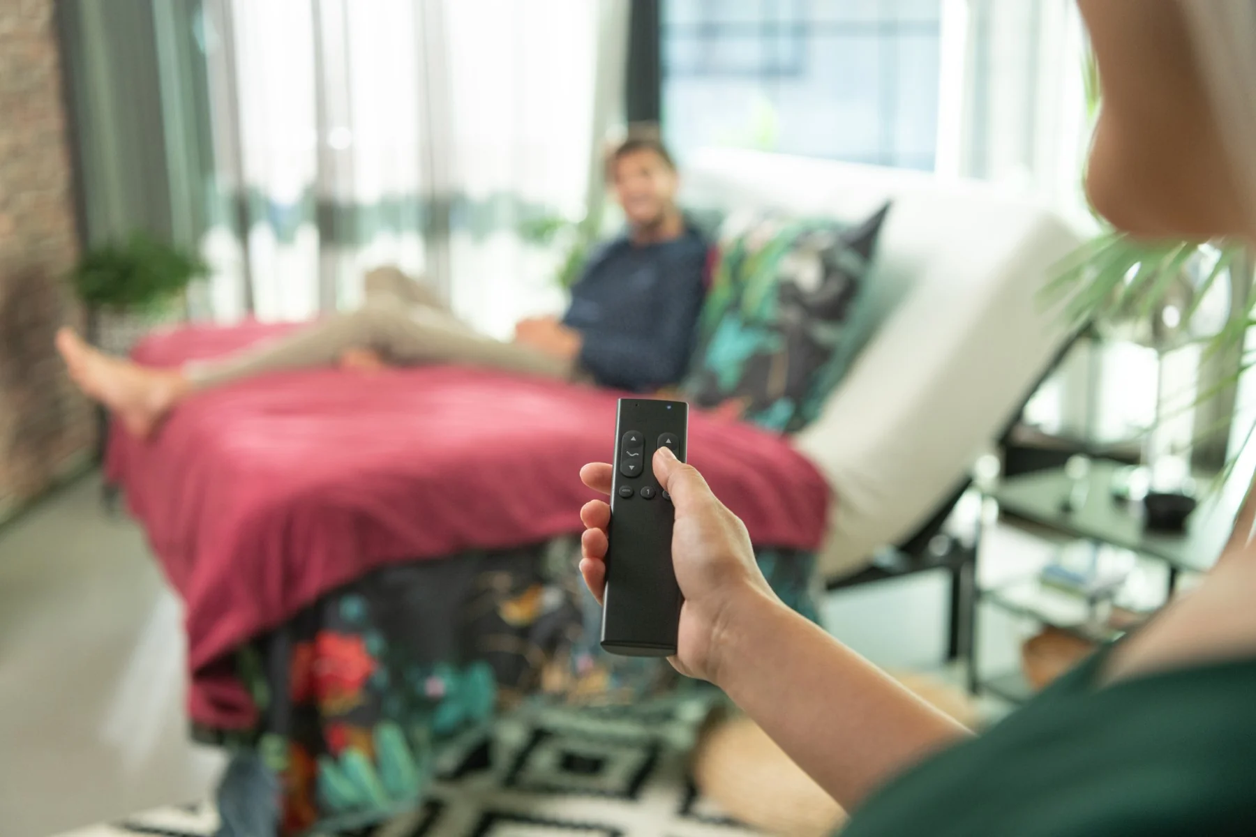 A person holding a remote control with another person lounging in the background on a bed.