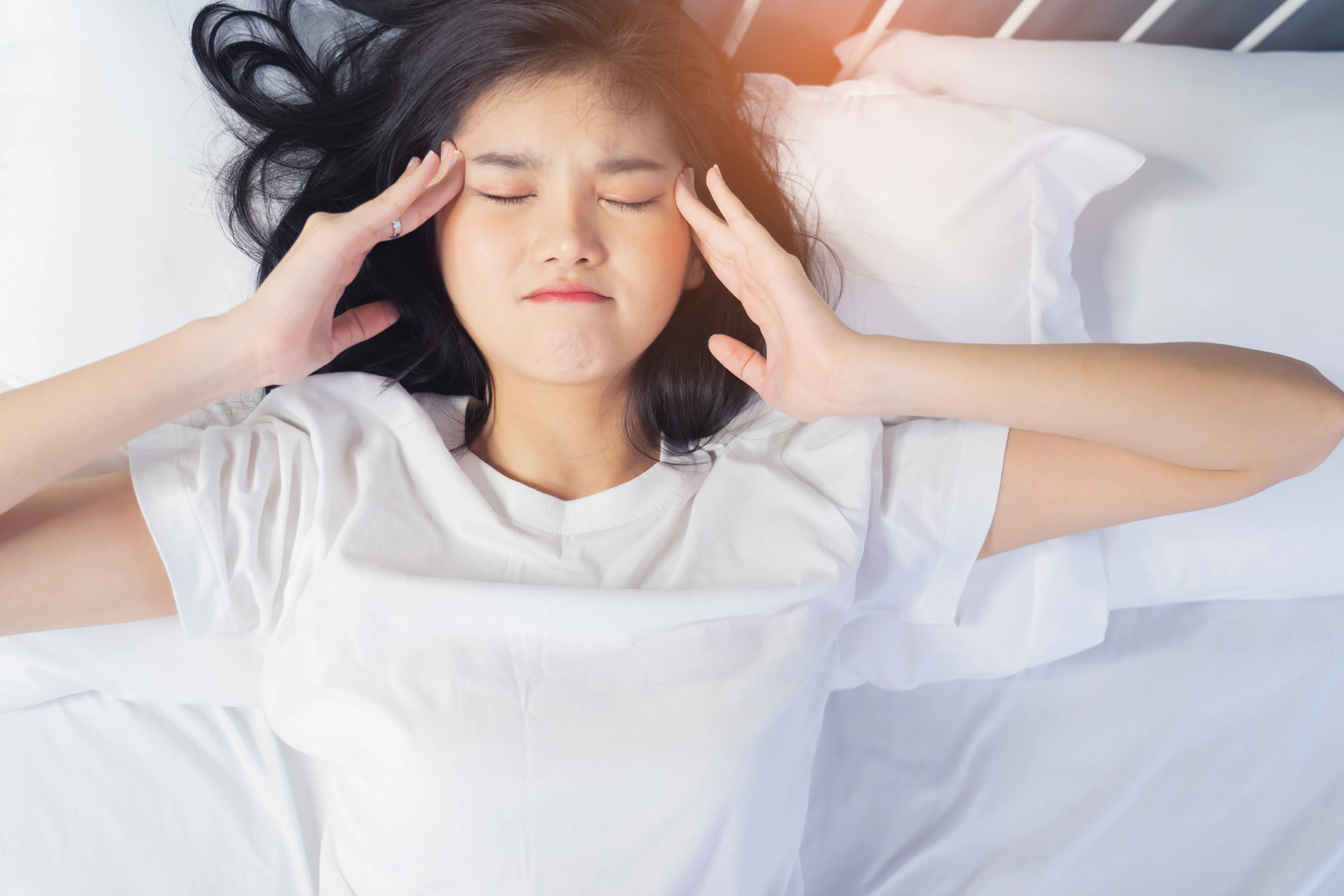 Young woman lying in bed with her eyes closed and hands on her temples, showing discomfort or dizziness, suggesting symptoms of vertigo while resting or waking up.