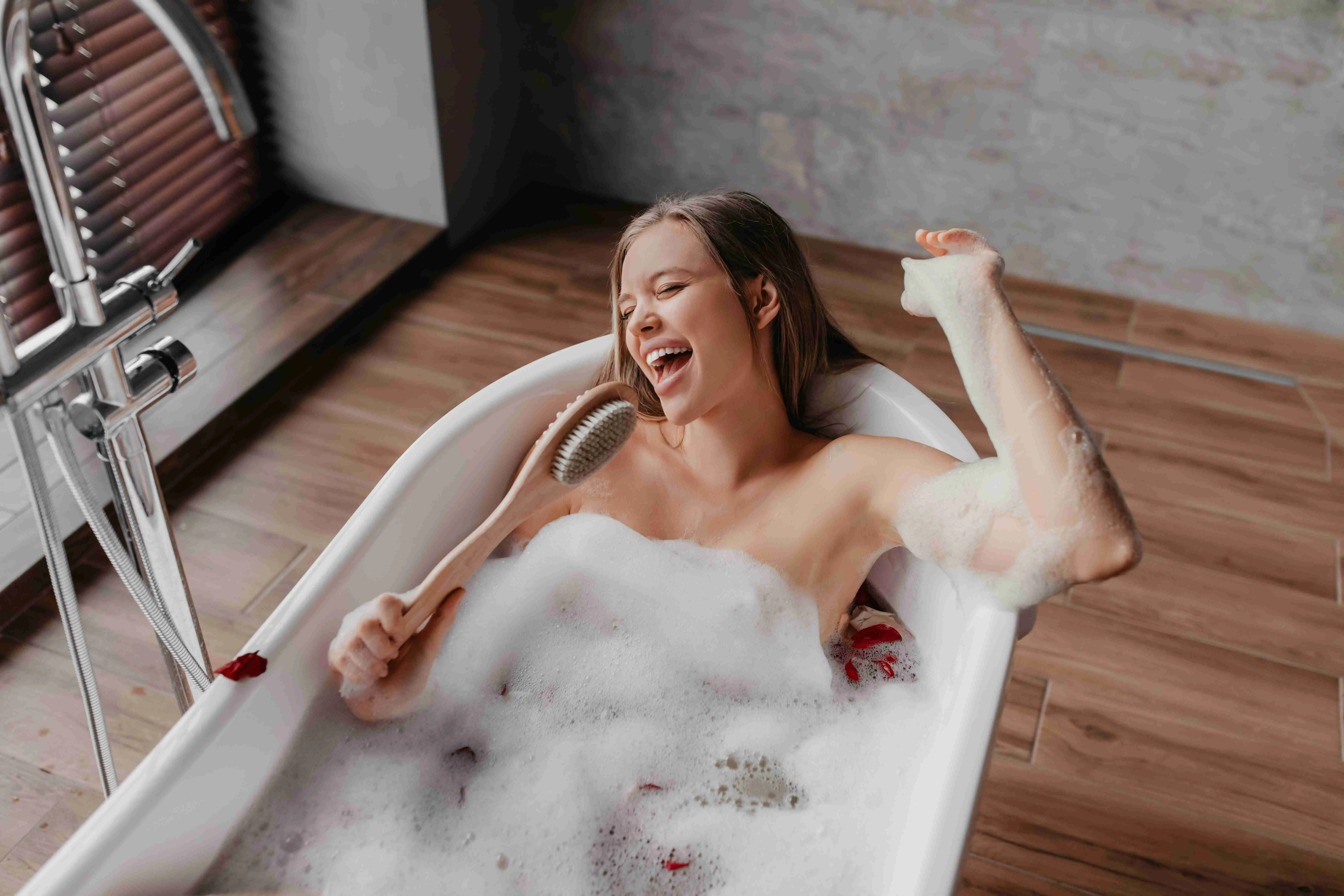 Happy lady singing song holding body brush as microphone, resting in foamy bathtub, giving way to talent and joy at free time in morning