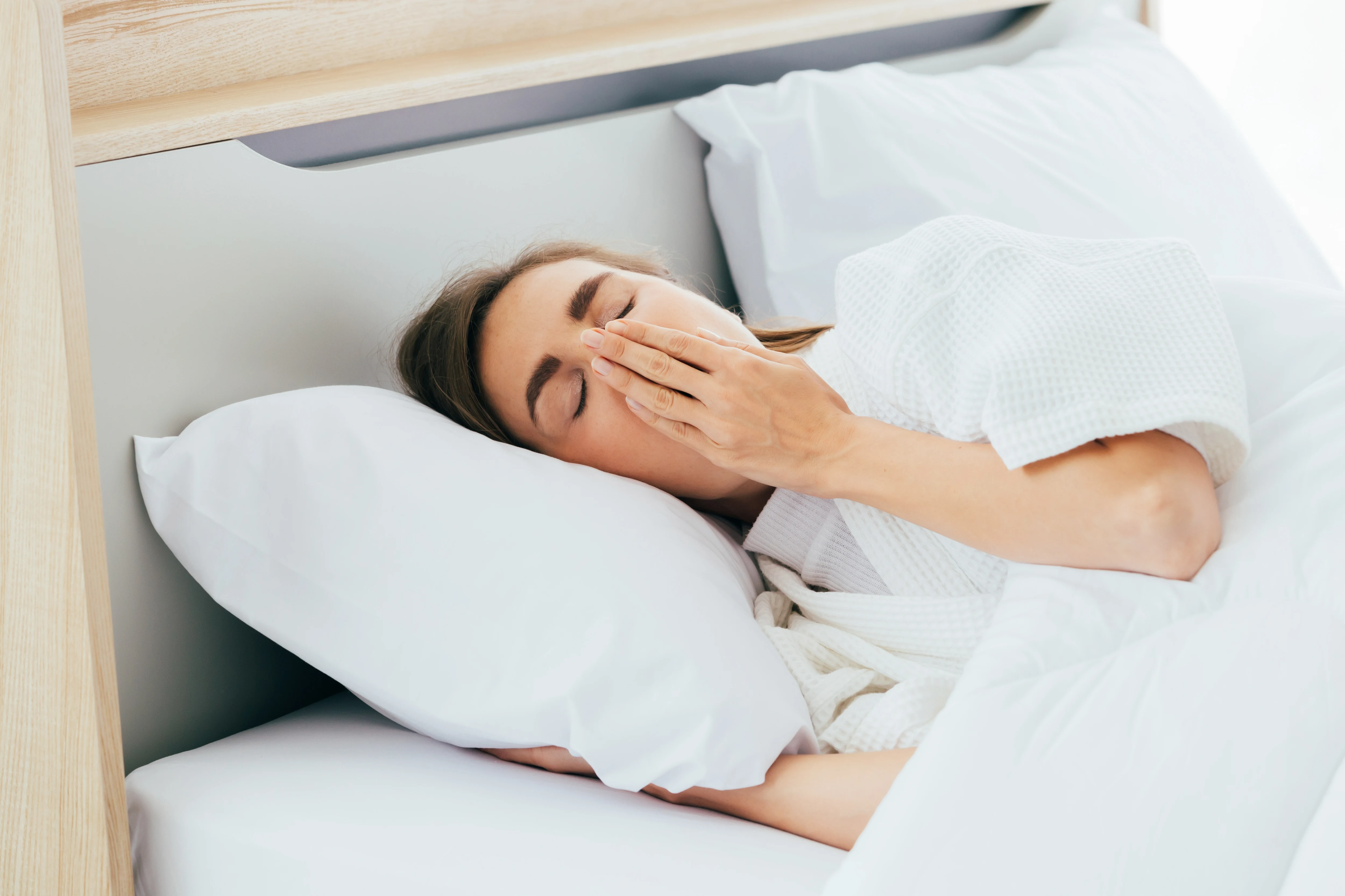 Sleepy Caucasian female with closed eyes covering mouth with hand and yawning while lying on comfortable bed in morning