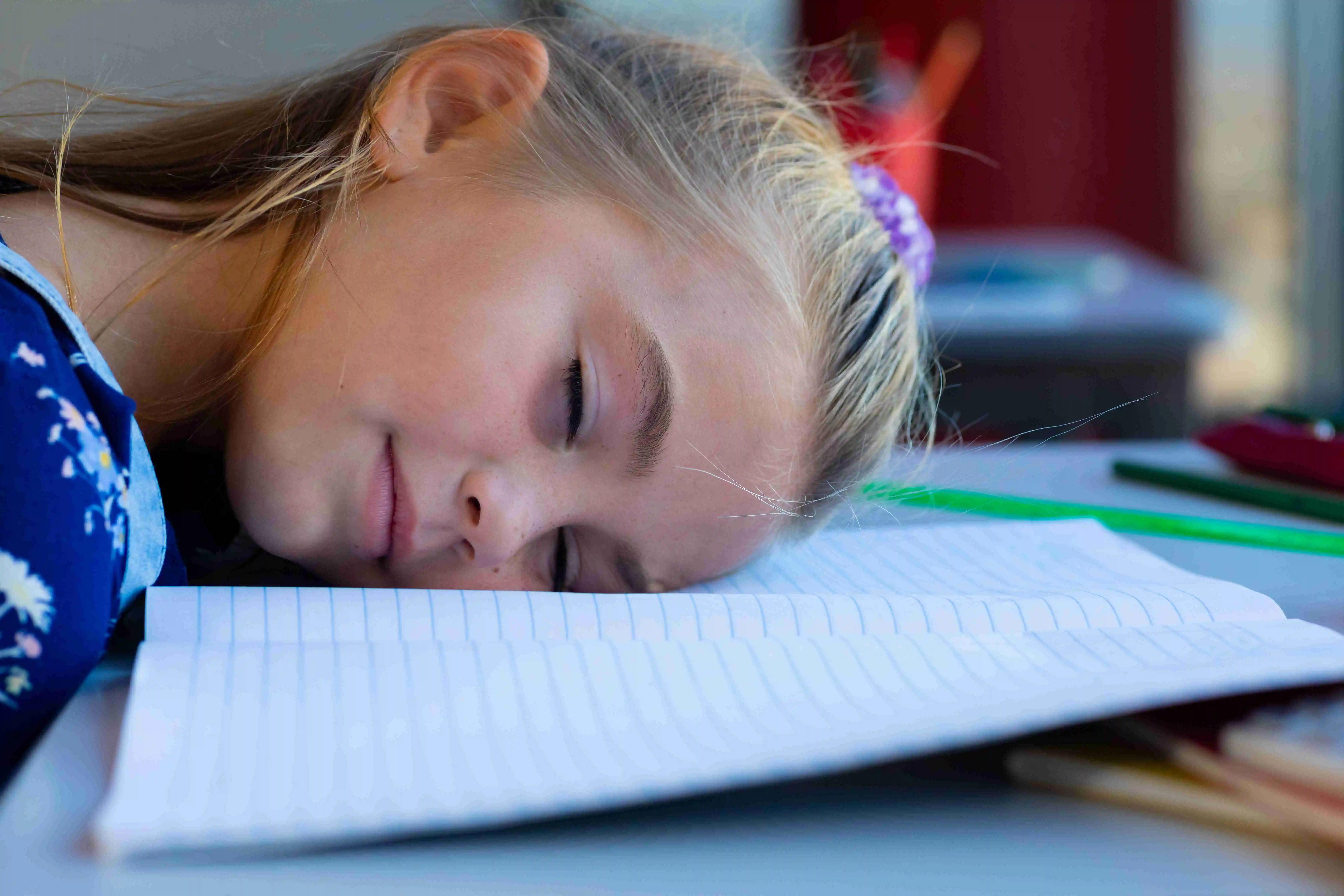 Tired caucasian schoolgirl sitting at desk and lying on books in classroom at elementary school