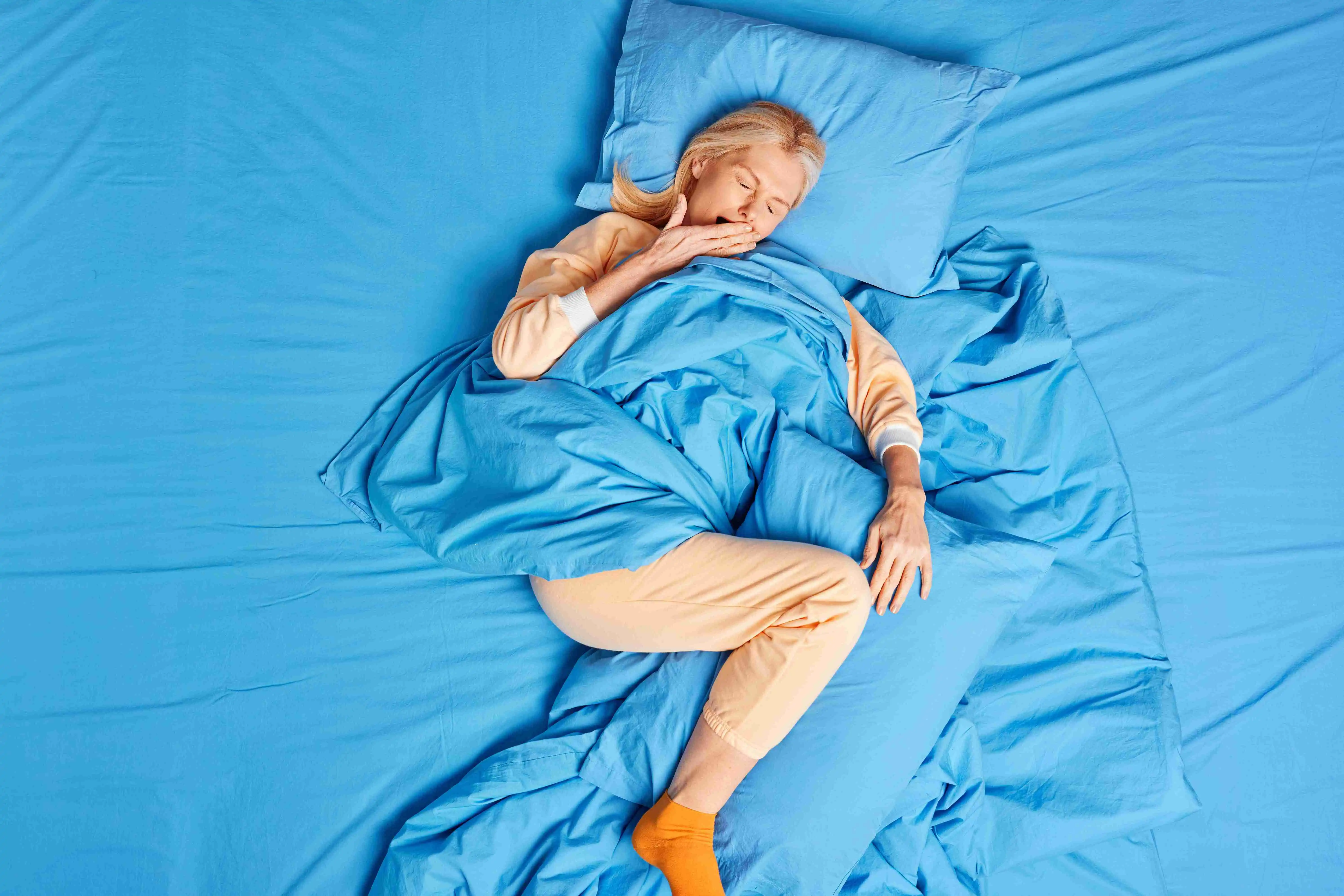 Top view of blonde forty years old European woman awakes early in morning yawns and covers mouth wears soft pajamas poses on bed had healthy sleep spends weekend at home