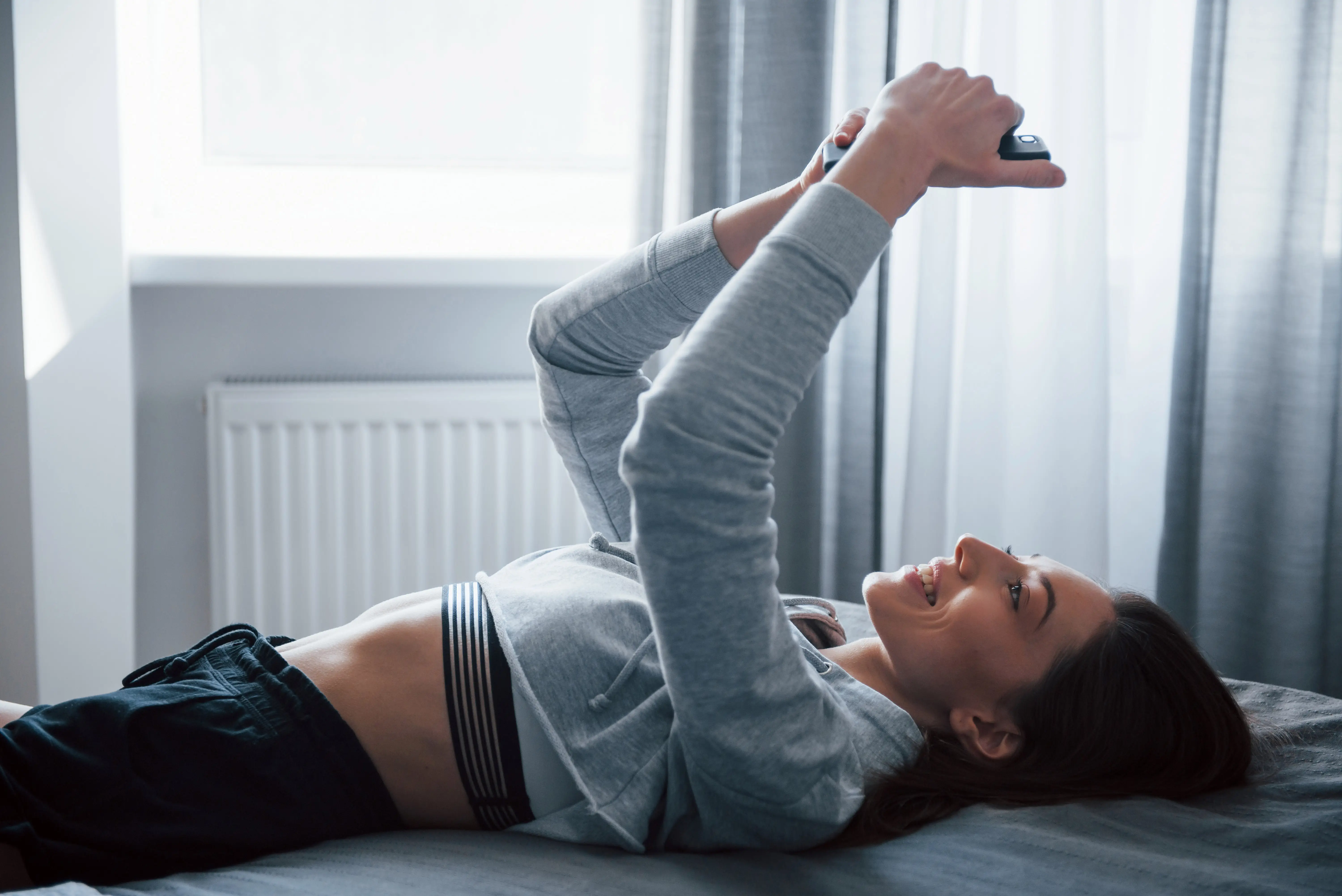 Side view of smiling girl taking selfie when lying on the bed near the window