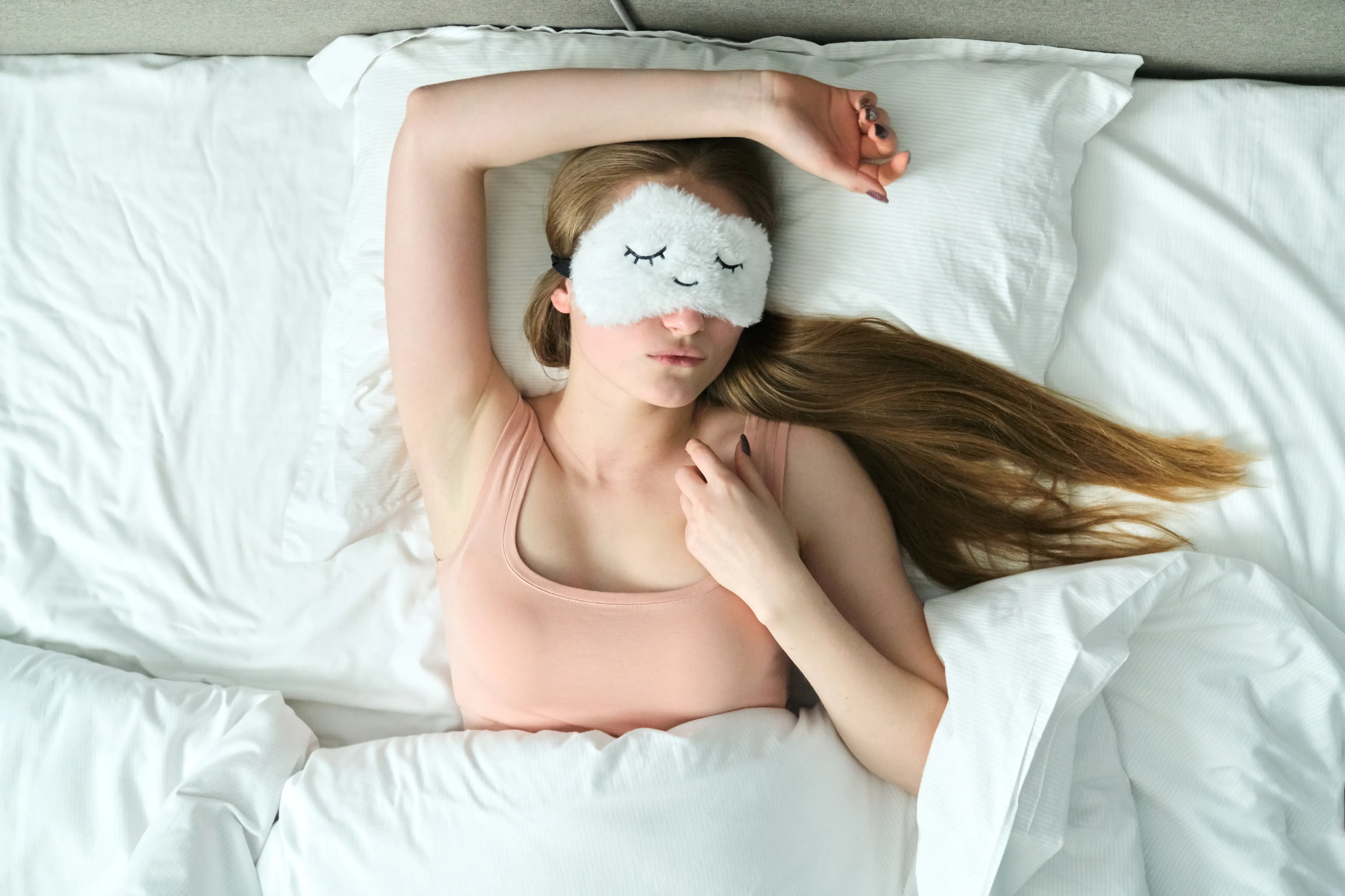 Young woman with long blonde hair with sleep mask in her eyes sleeping in white bed, top view