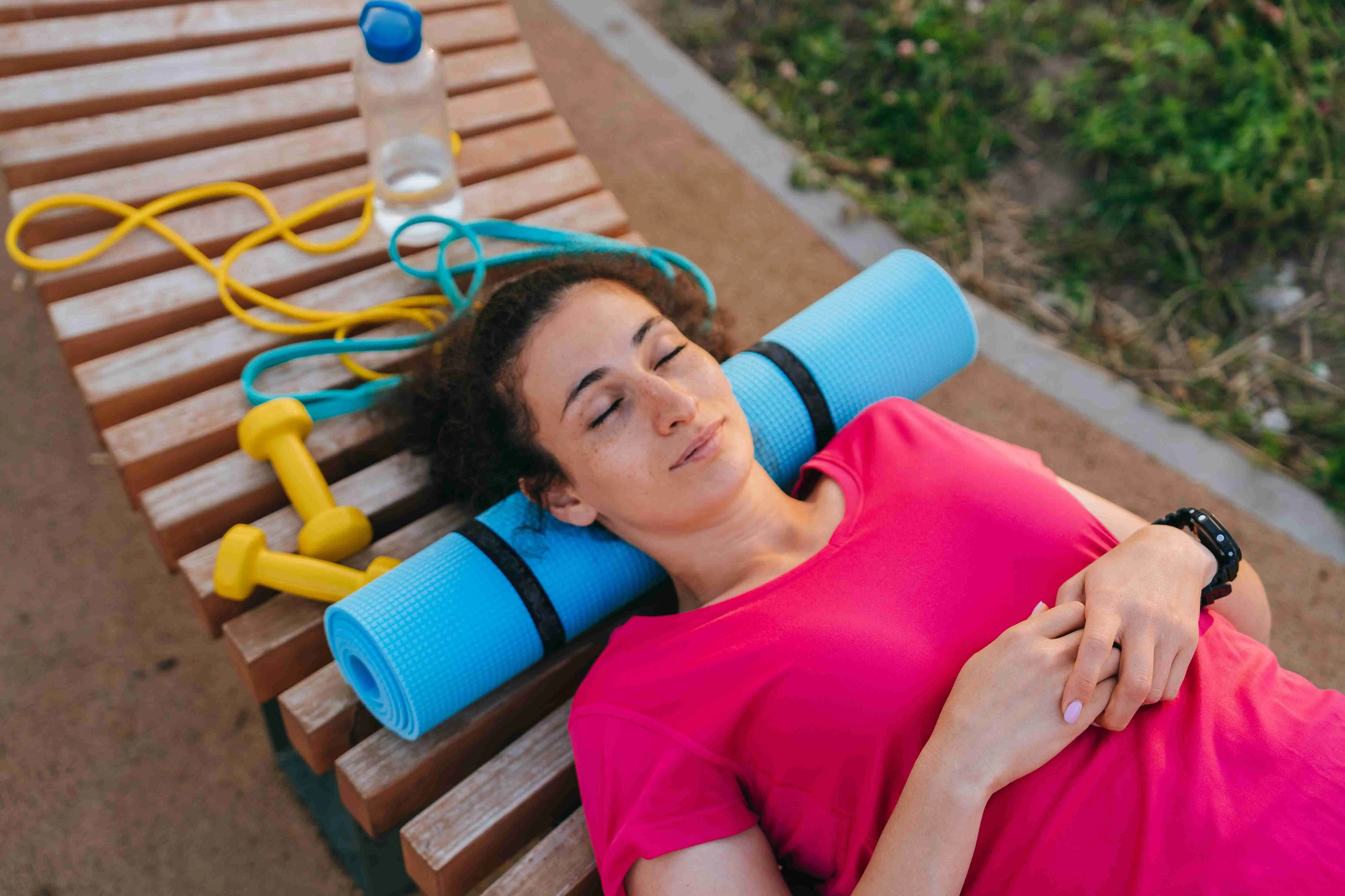 Relaxed woman lying on a bench with yoga mat and exercise equipment after workout, enjoying rest