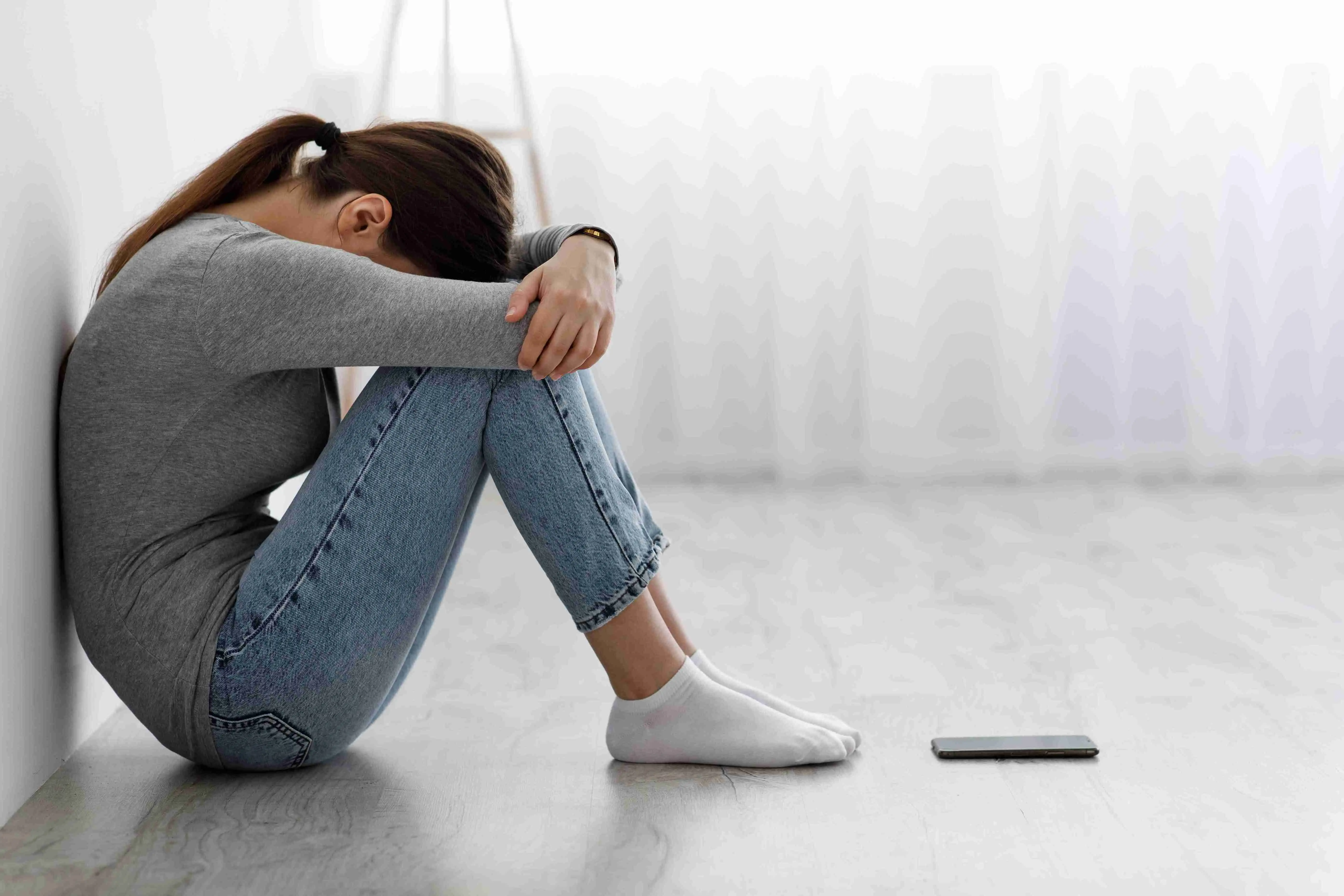 Sad unhappy depressed millennial Caucasian female sitting on floor with phone, covering her face