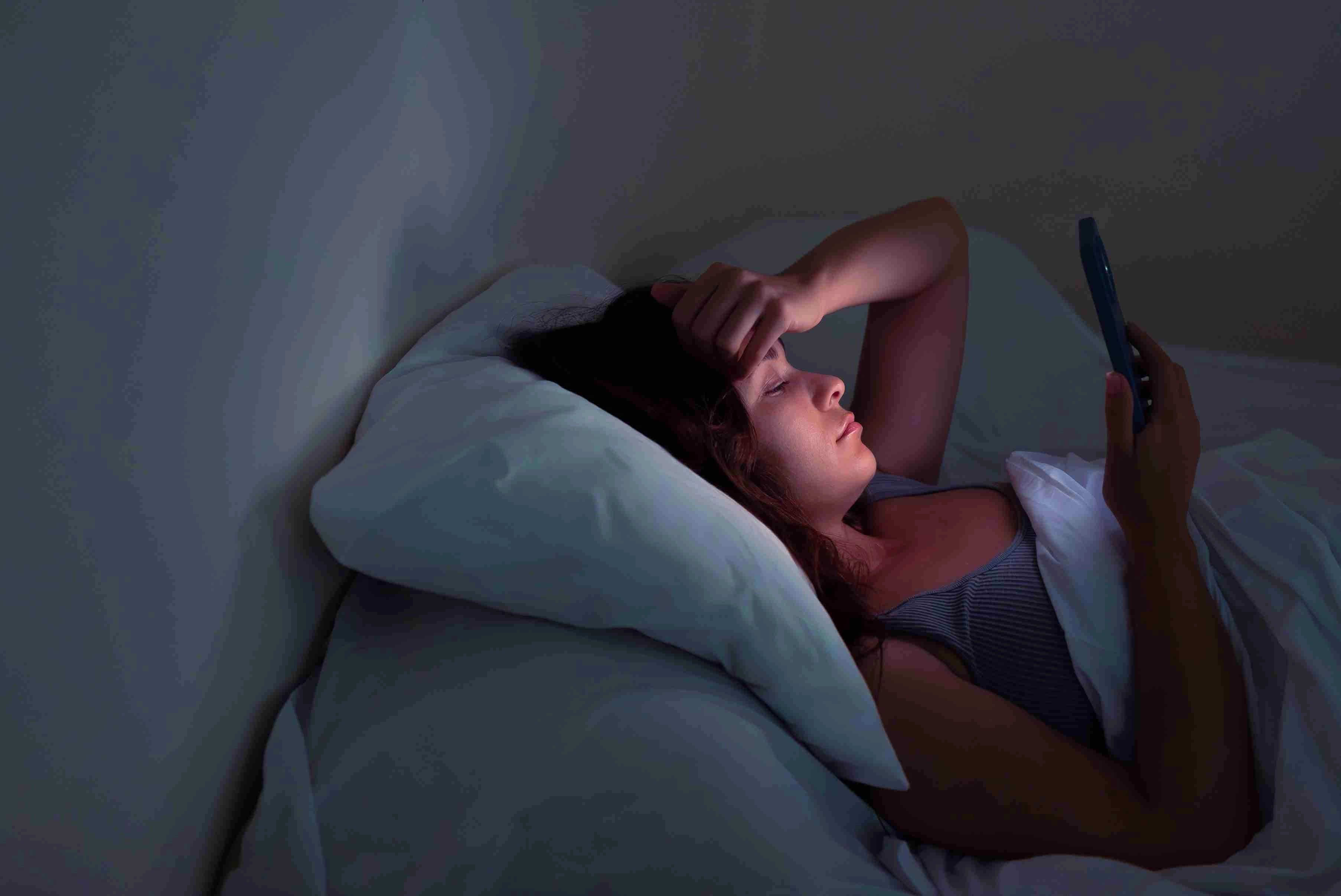 A young woman, grappling with insomnia, finds comfort in her smartphone's warm illumination