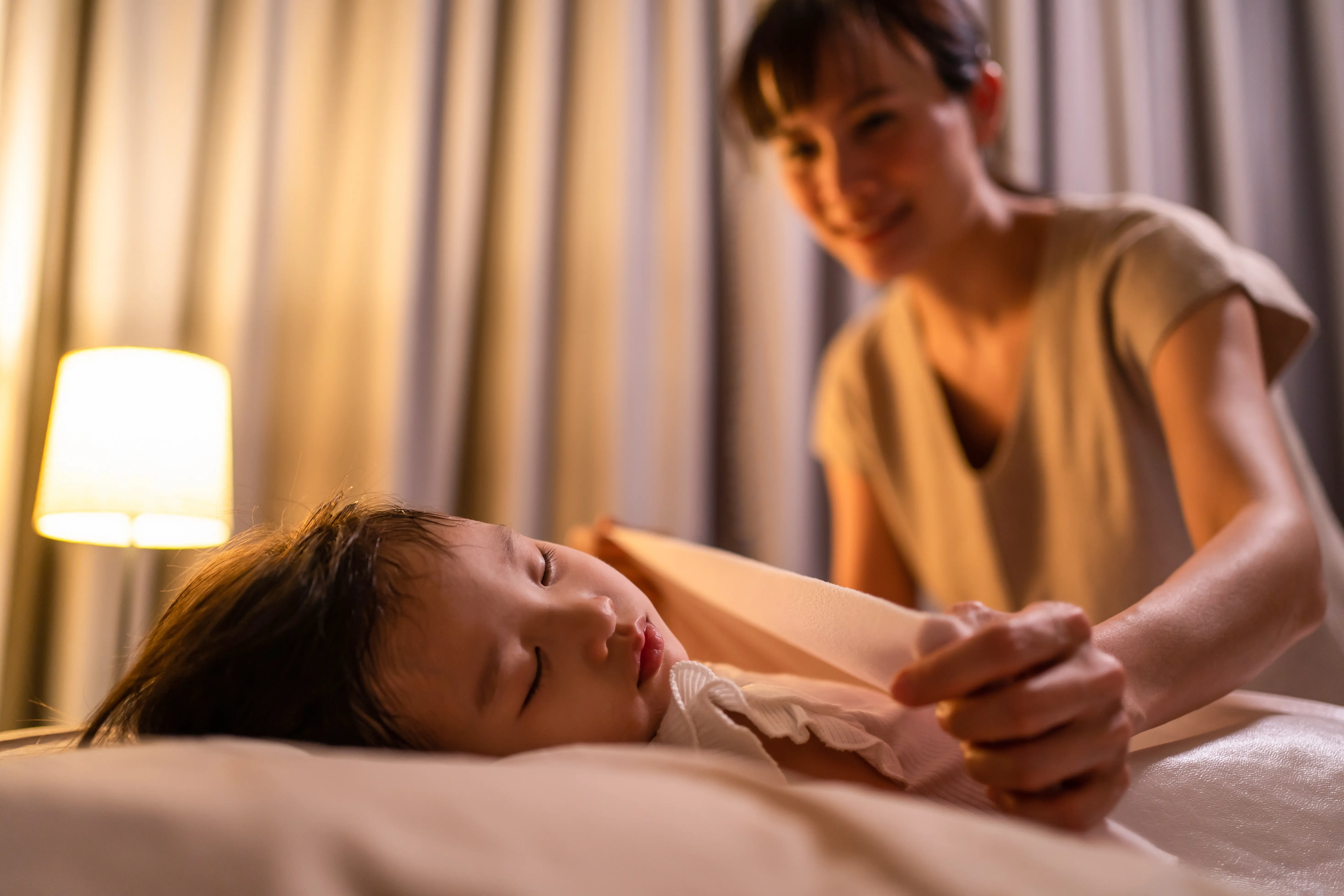 Asian mother put blanket on sleeping little small girl daughter on bed in bedroom at night.